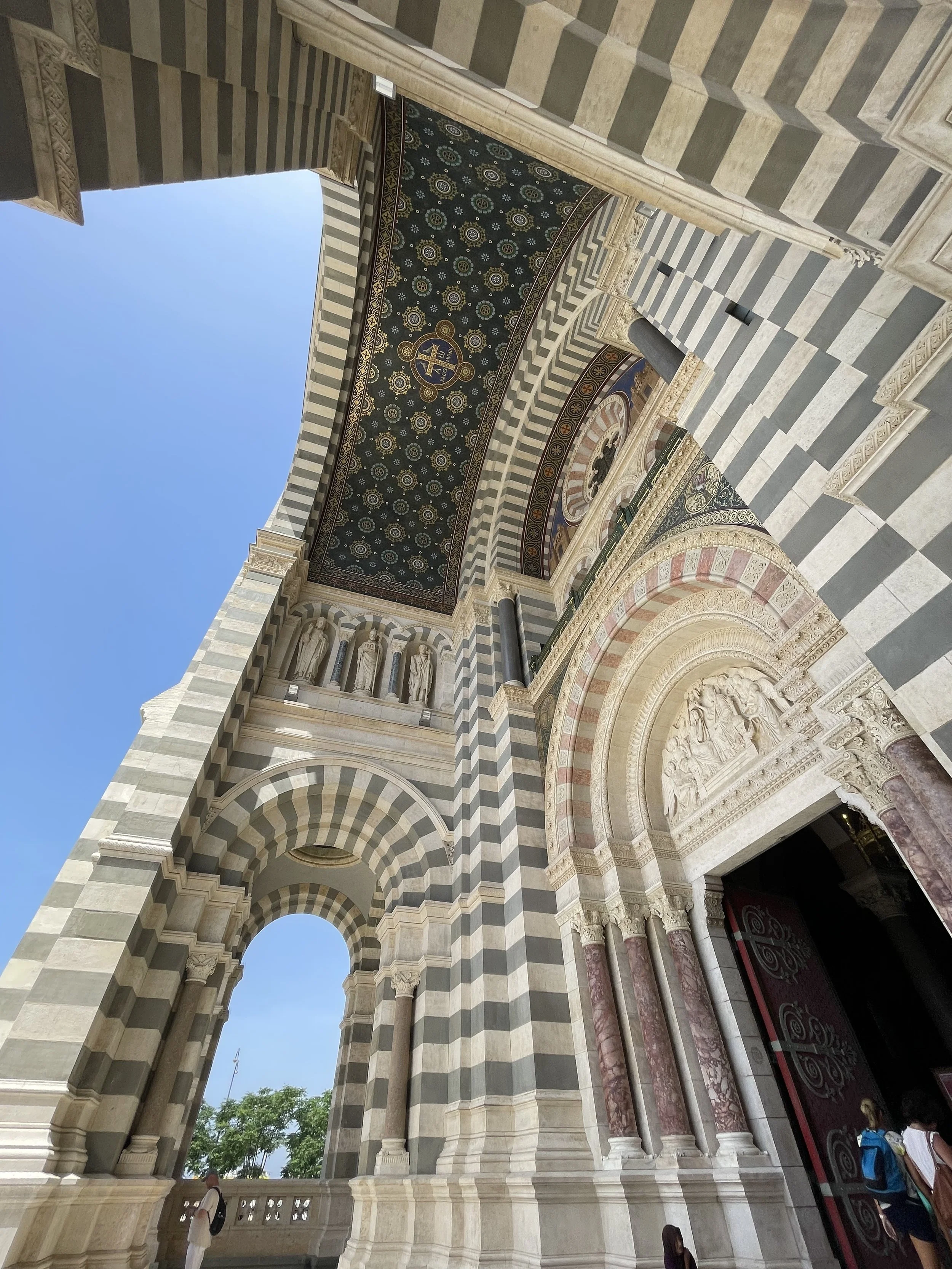Exterior view of a historic cathedral with striped black and white marble columns, arches, and intricate stone carvings, under a clear blue sky.