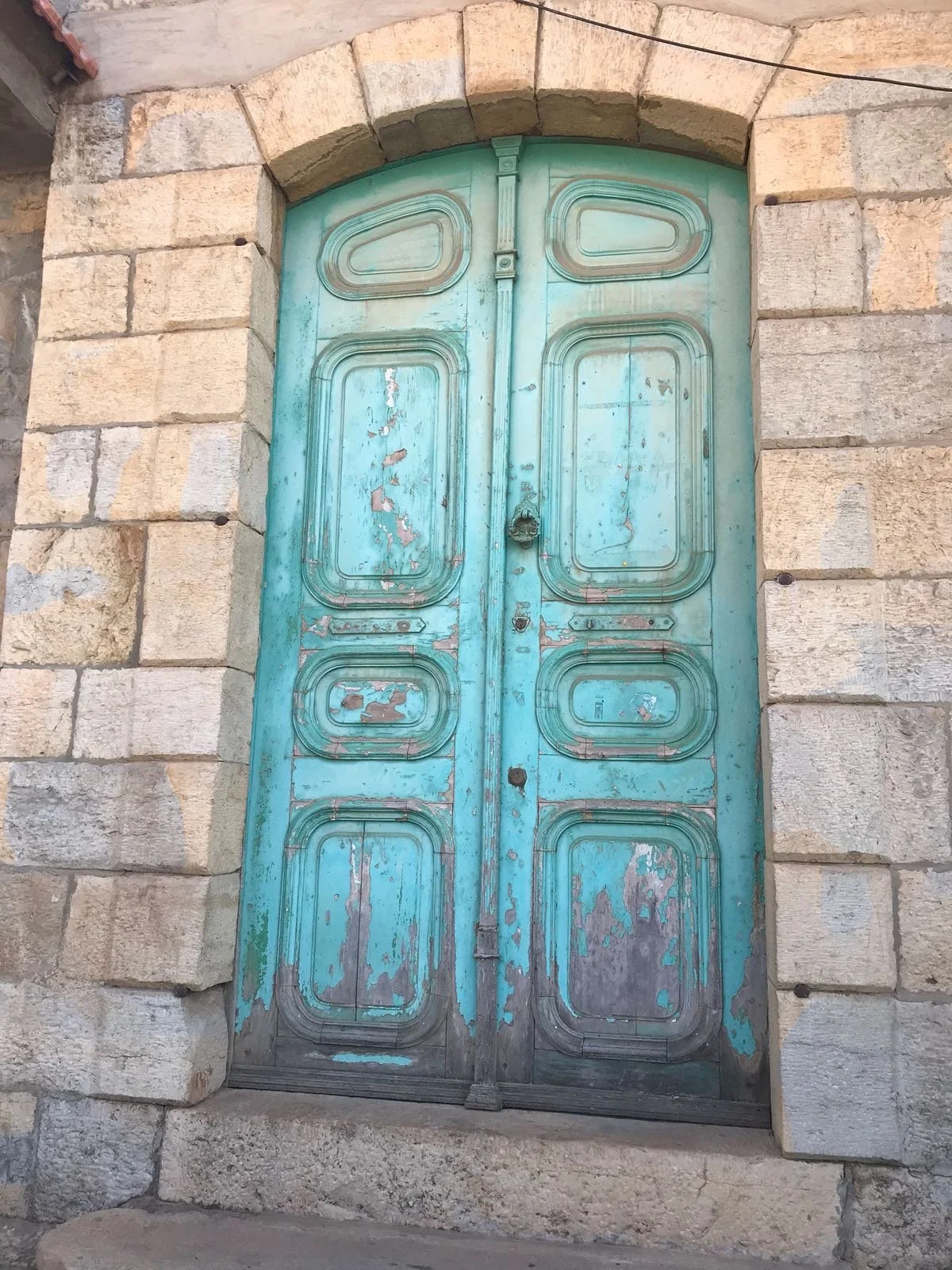 A worn, turquoise-painted wooden door with decorative panels, set within a stone archway, on a stone wall.