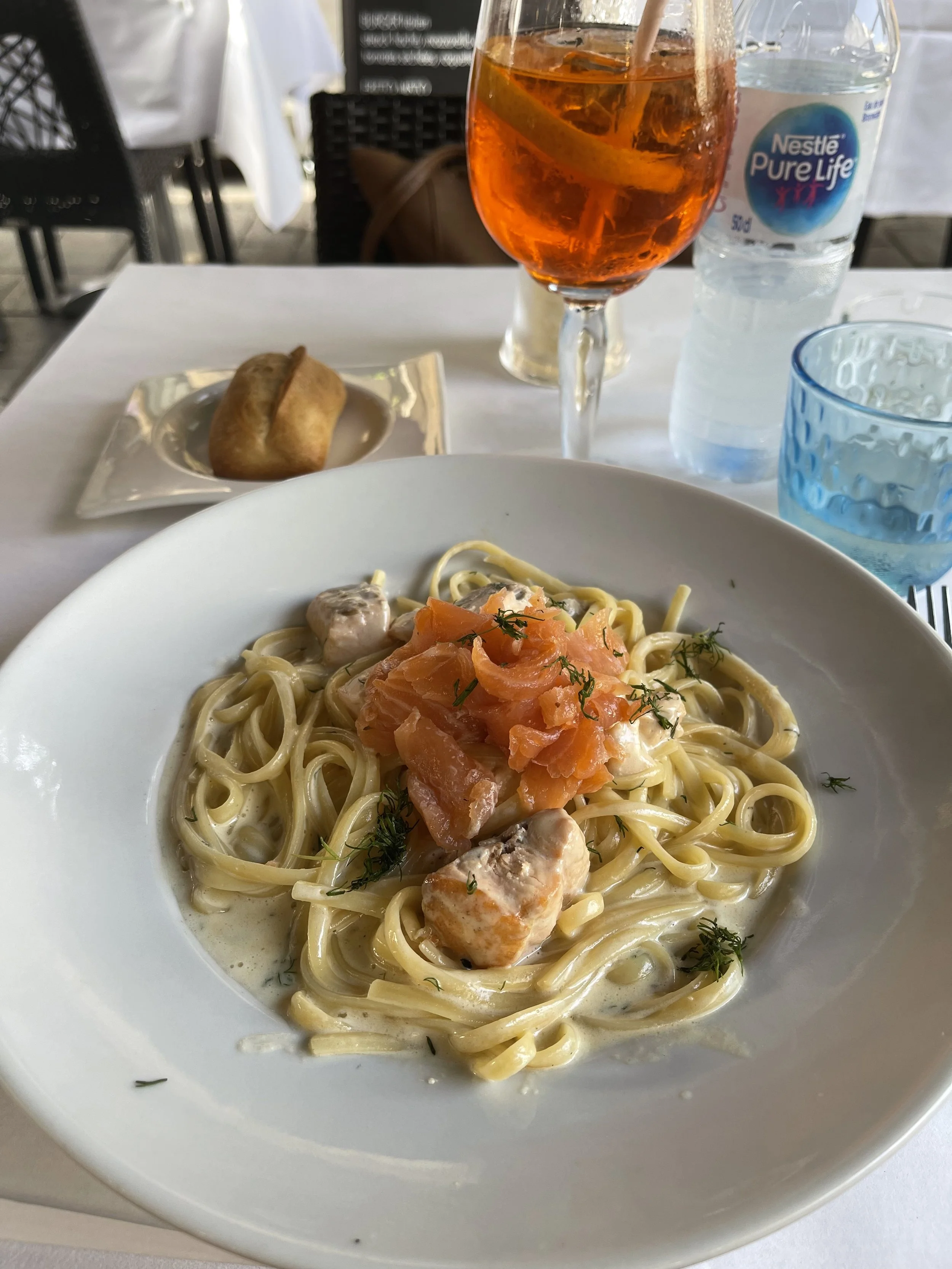A white plate of fettuccine pasta with salmon pieces and a creamy sauce, garnished with herbs, on a table in a restaurant setting. In the background, there is a glass of orange-colored drink with ice, a bottle of water, a small bread roll on a silver