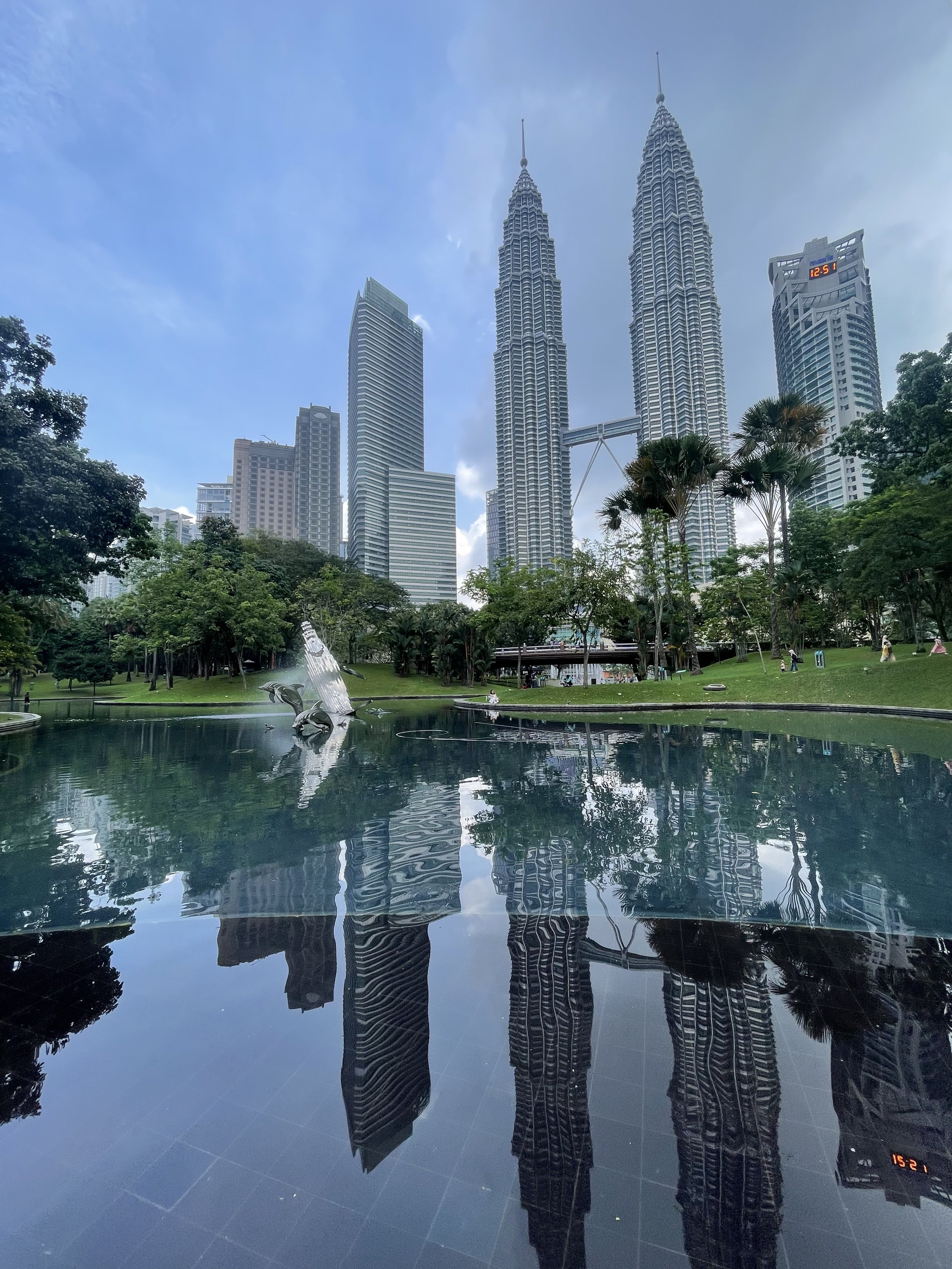 A cityscape featuring the Petronas Towers and other high-rise buildings reflected on a body of water in a park with trees and sculptures.
