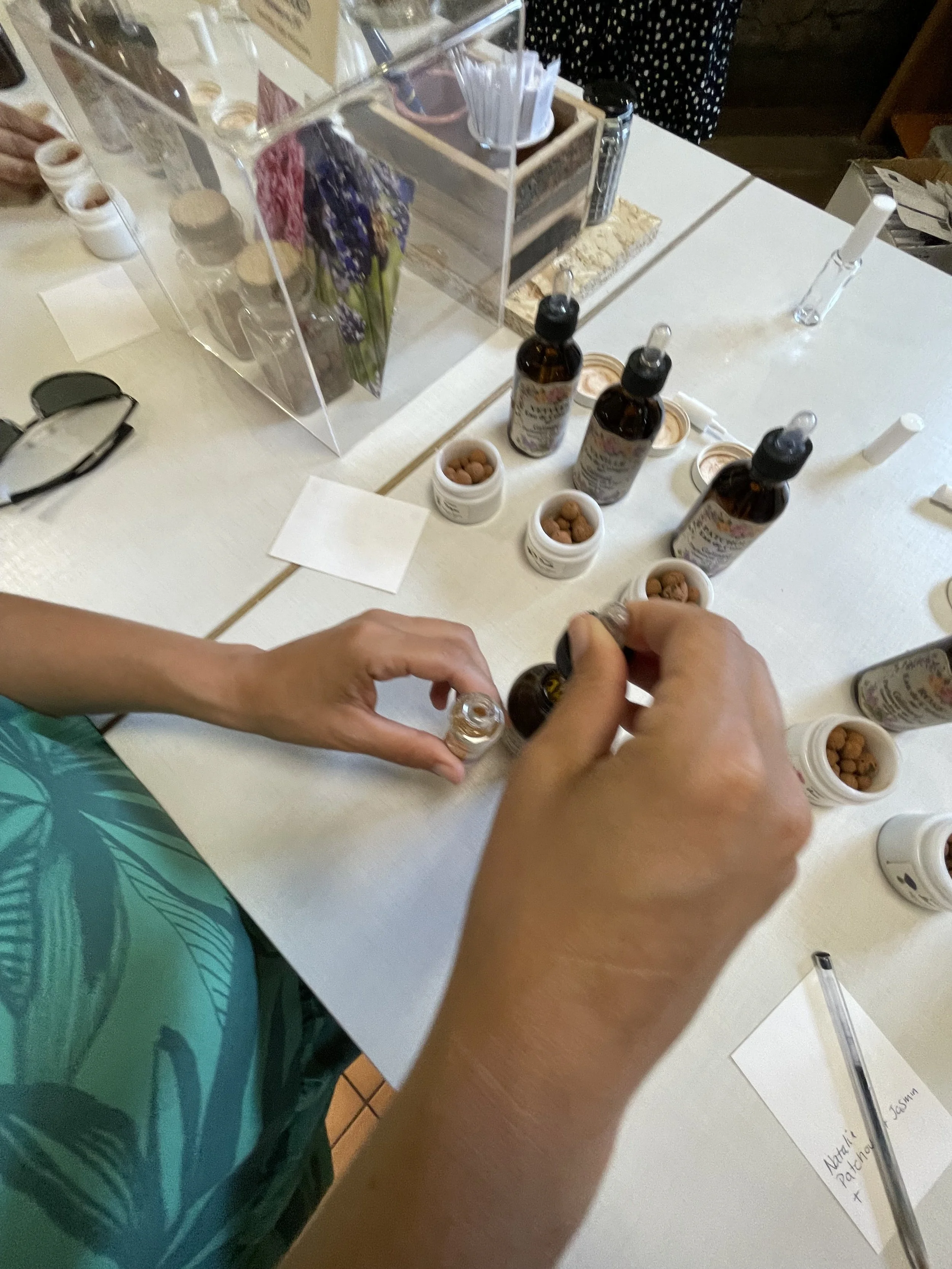Person filling small glass bottles with essential oil in a workshop setting with bottles, jars, pens, and notes on a table.