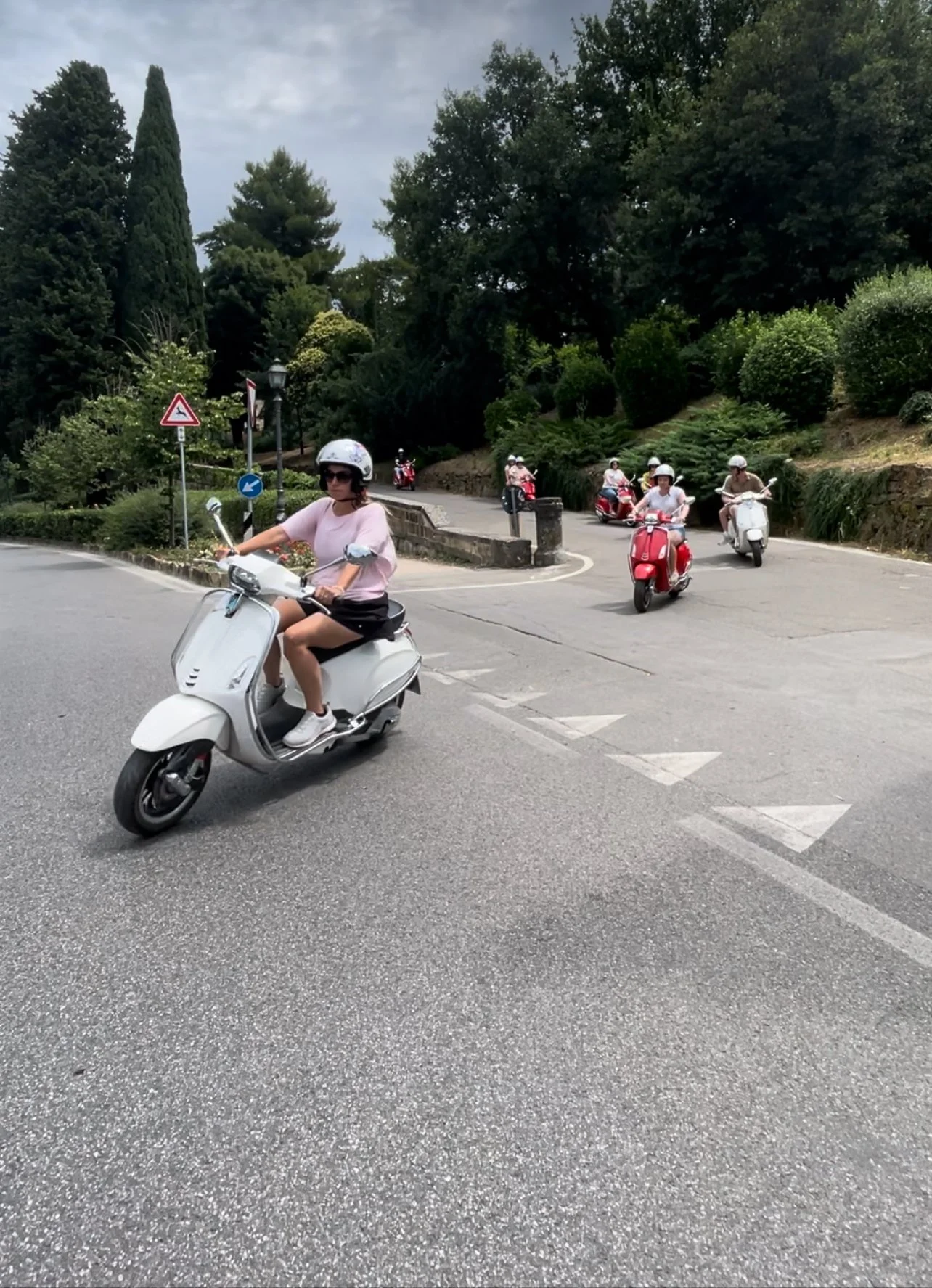 Several women riding scooters on a curved road surrounded by lush green trees and bushes, with traffic signs nearby.