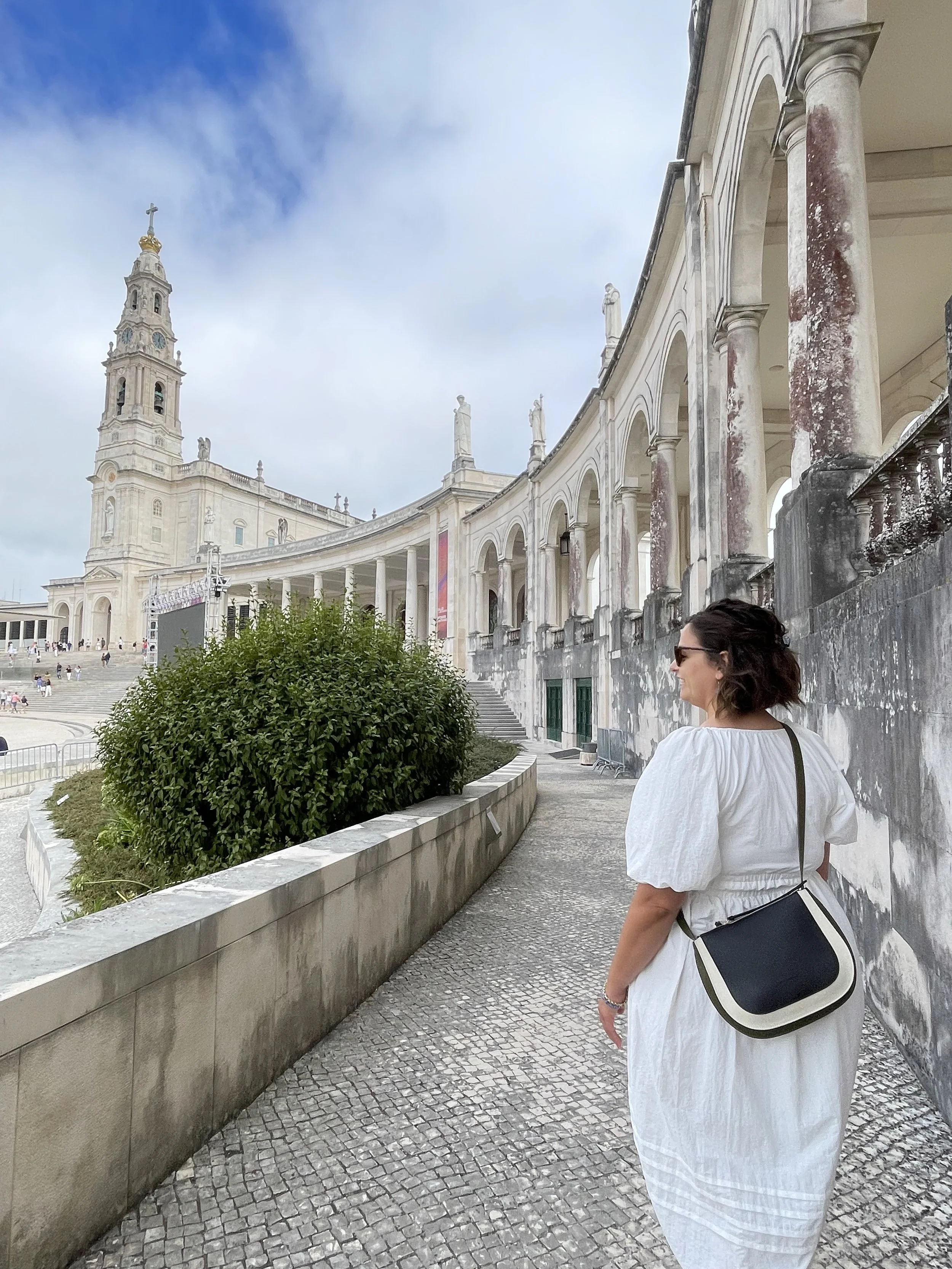 A woman in a white dress with short sleeves and sunglasses walking along a curved, stone-paved outdoor corridor of a historical building with white columns and statues, with a church or cathedral in the background under a partly cloudy sky.