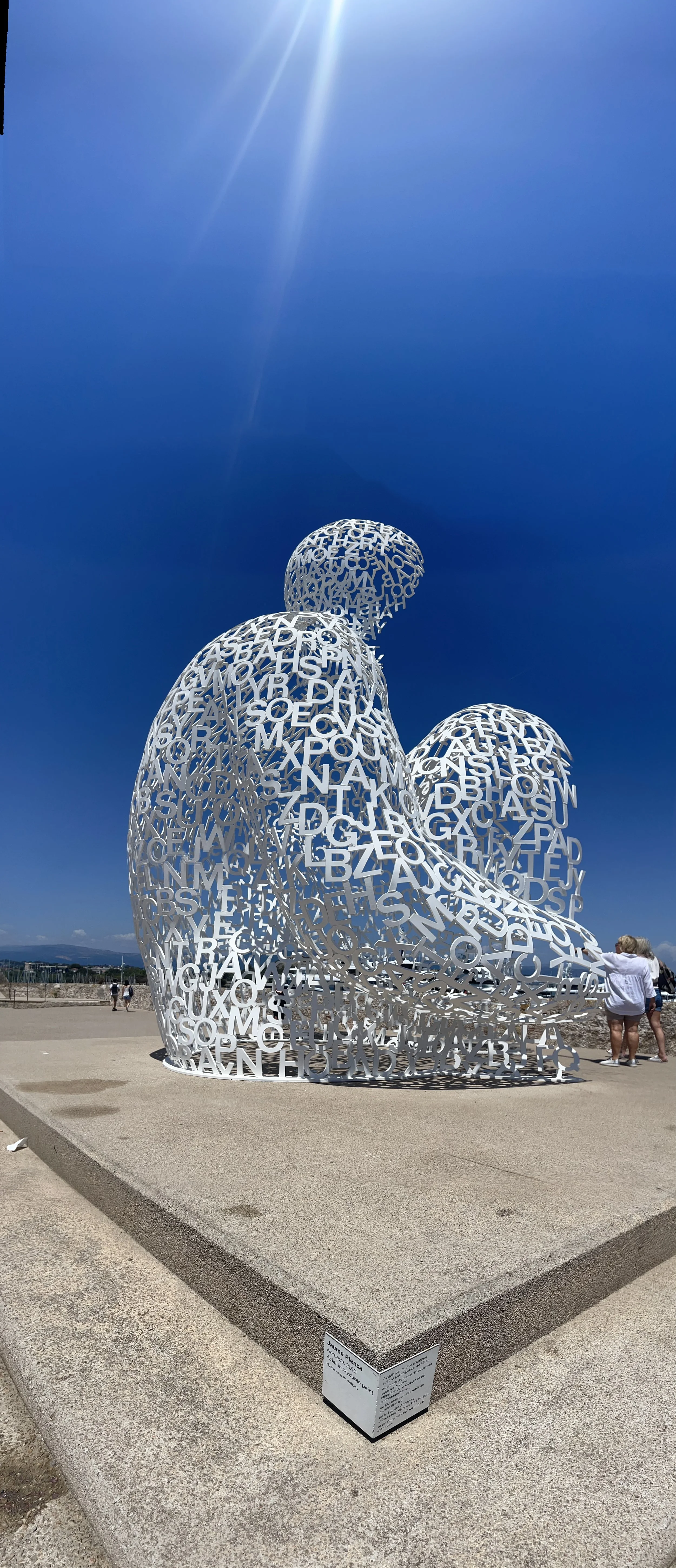 Large outdoor sculpture of a heart made of white metal letters, located on a concrete platform with a clear blue sky overhead.
