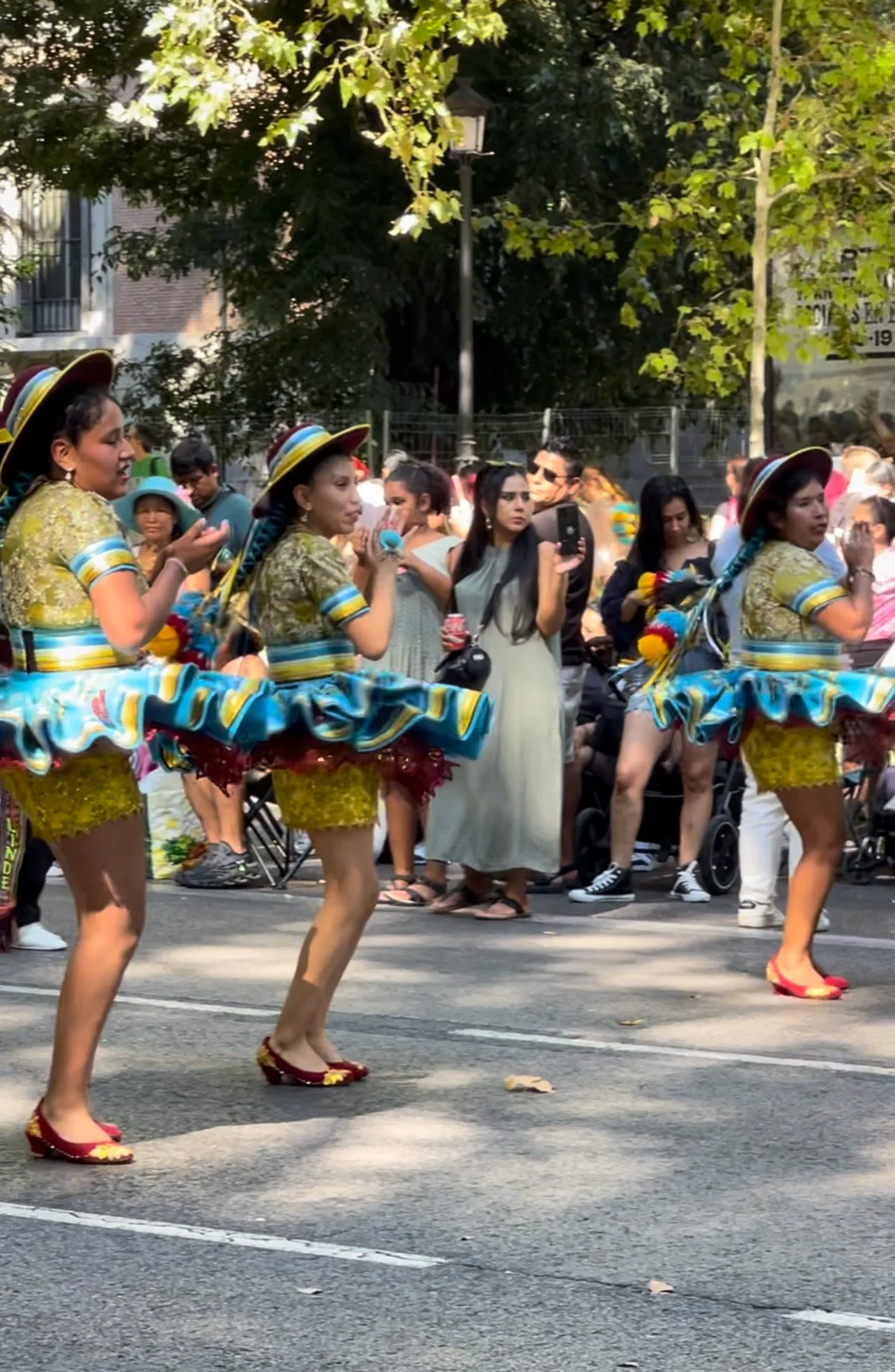 Women dressed in colorful traditional costumes, dancing in a parade on a city street, with a crowd of spectators watching and taking photos.