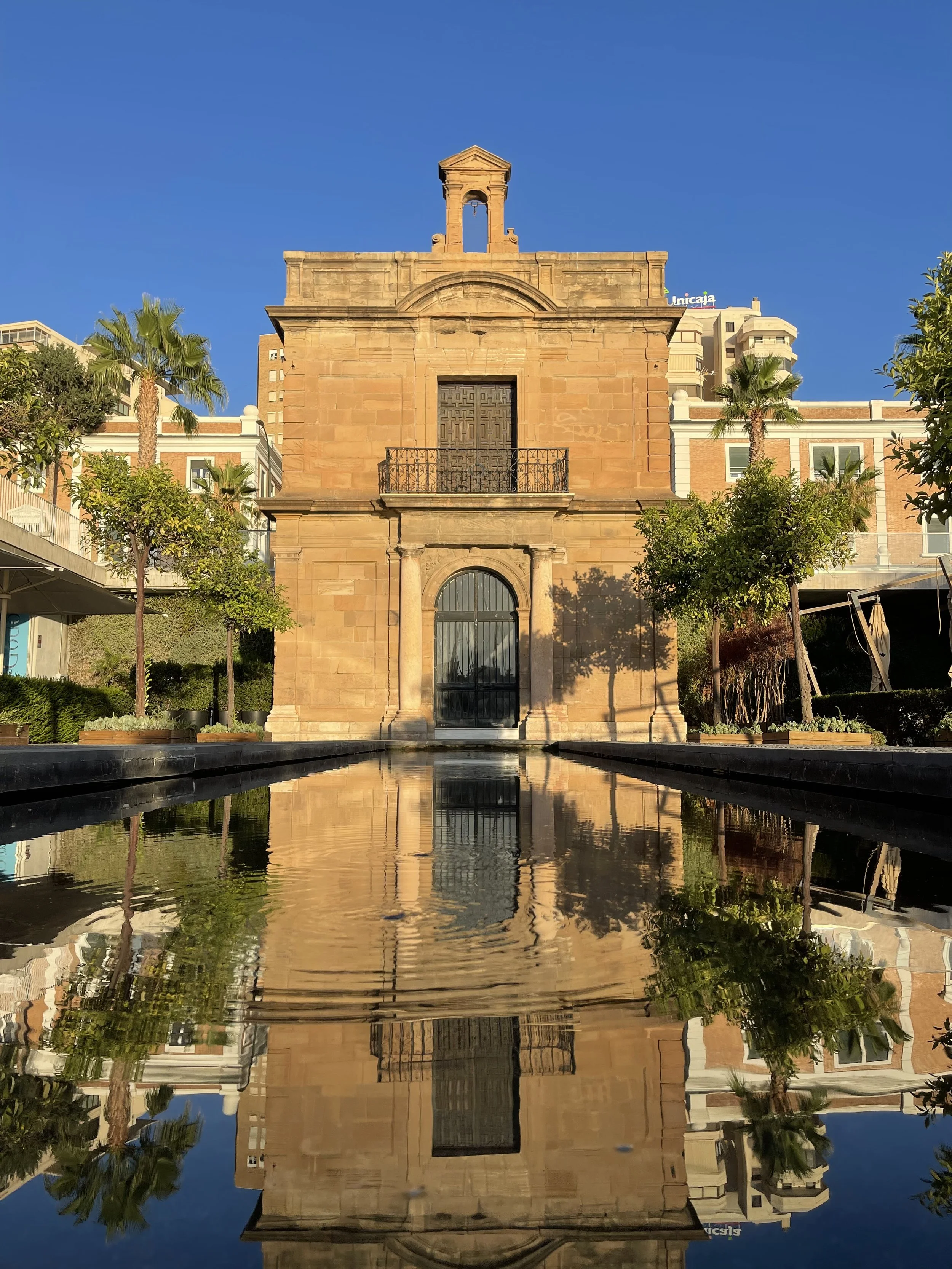 A historic stone building with a bell tower, reflected in a pool of water, surrounded by trees under a clear blue sky.