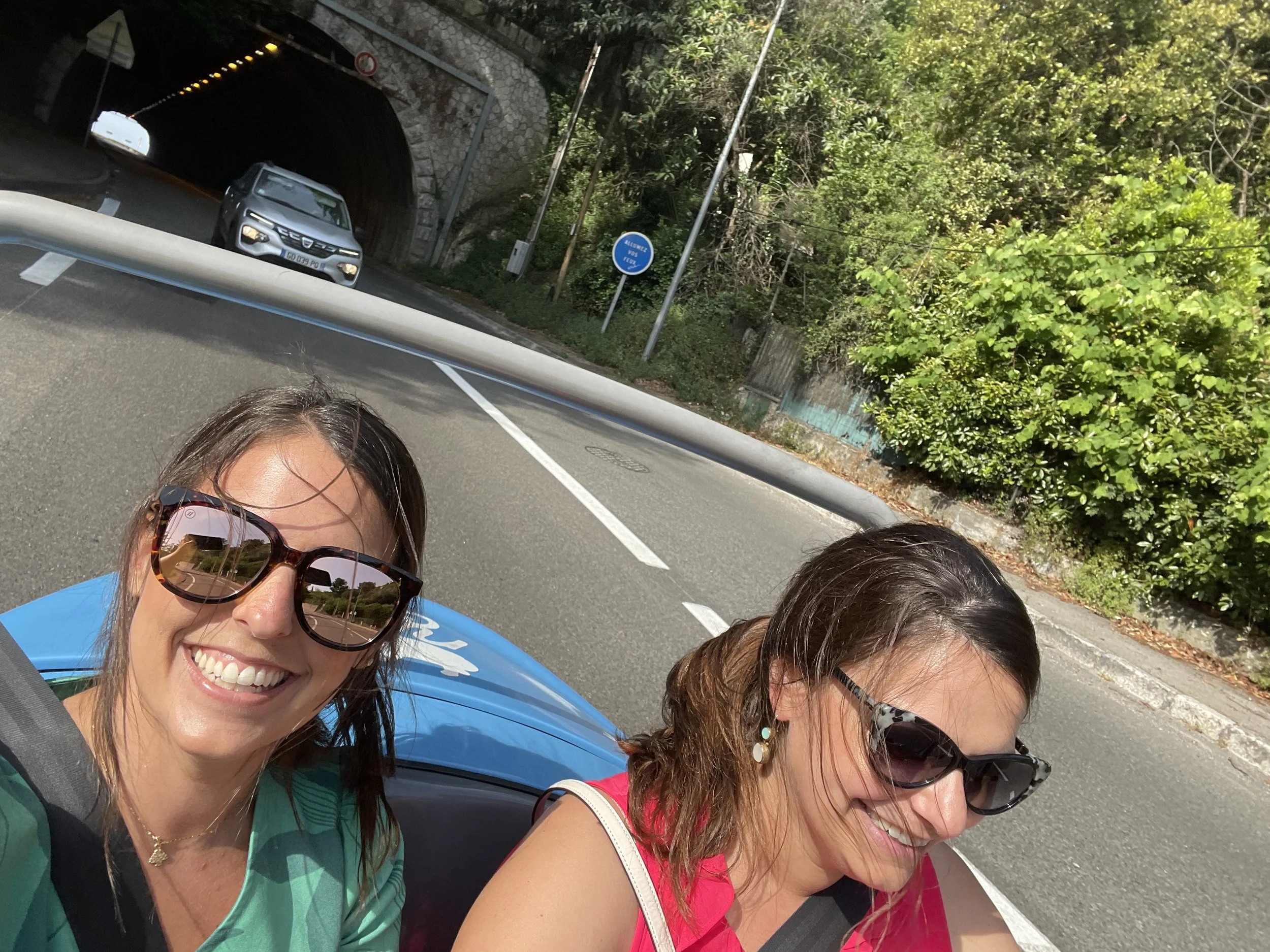 Two women are taking a selfie while riding in an open-top vehicle, with a scenic road and green trees in the background. Both are wearing sunglasses and smiling.