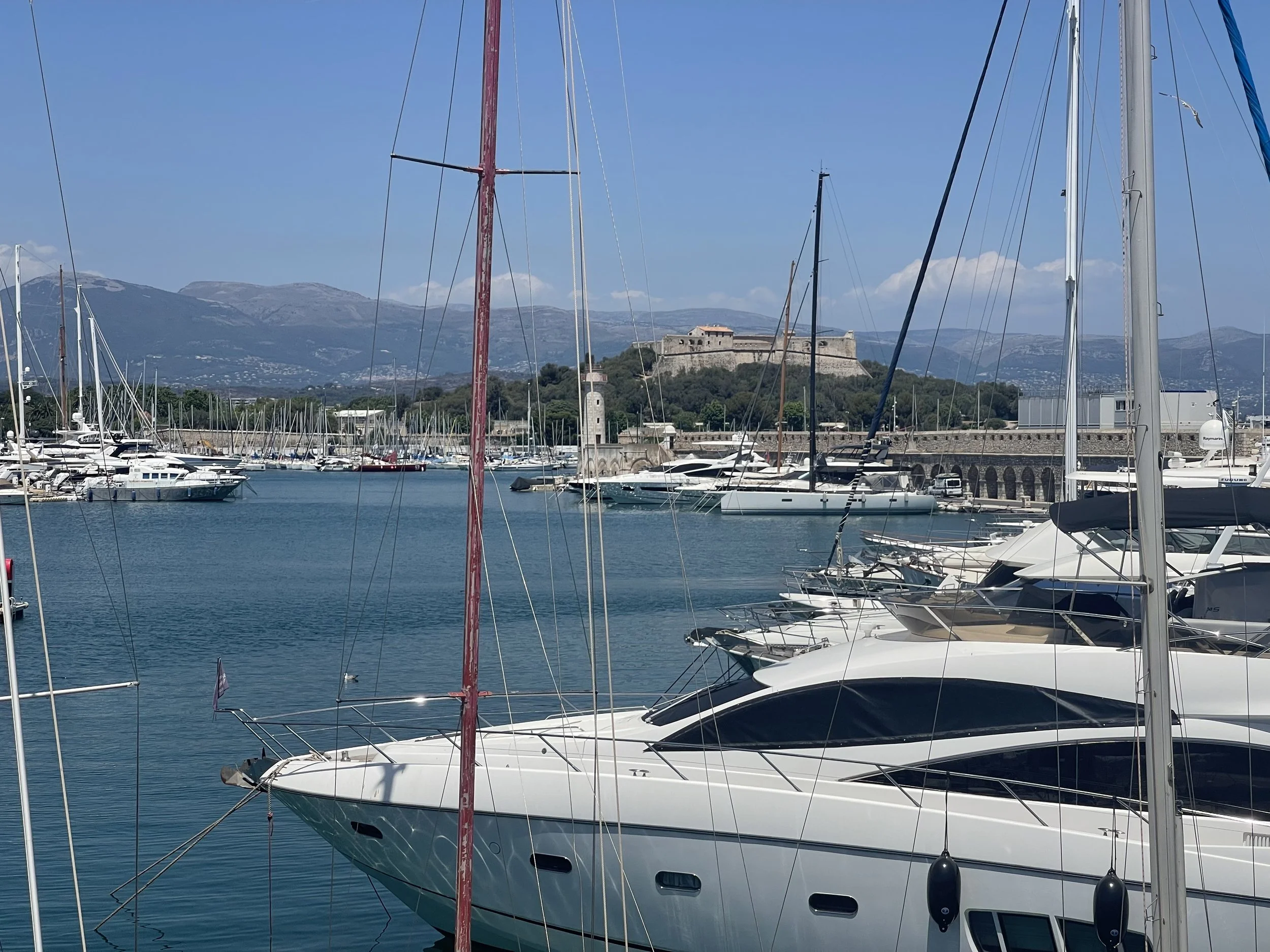 Glamorous marina with luxury yachts anchored, a historic fortress on a hill, and distant mountains in the background under a clear blue sky.