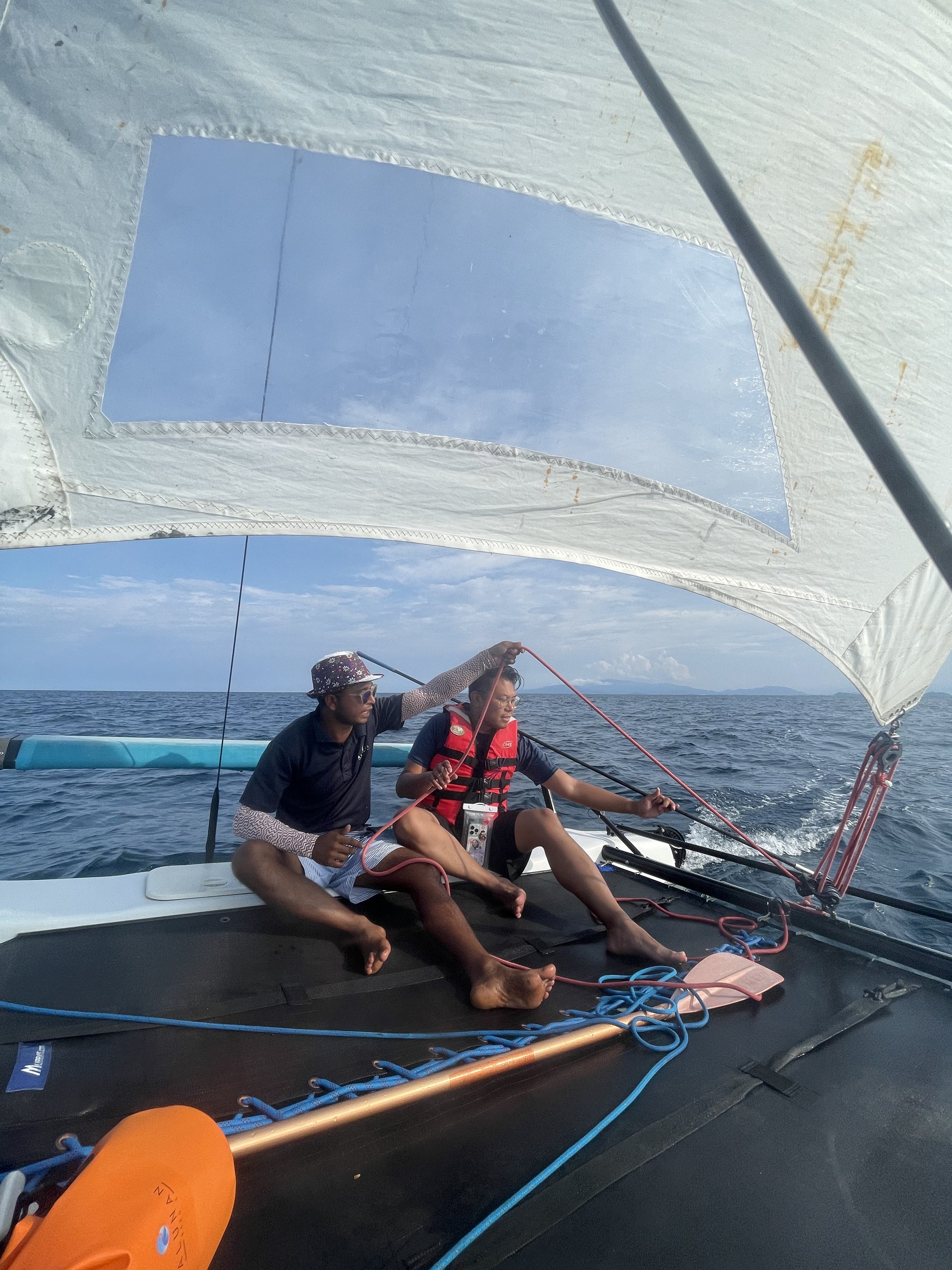 Two people sit on the deck of a sailboat, with the ocean and sky visible in the background. One person is adjusting the sail while the other looks on. They are both dressed casually and wearing life jackets.