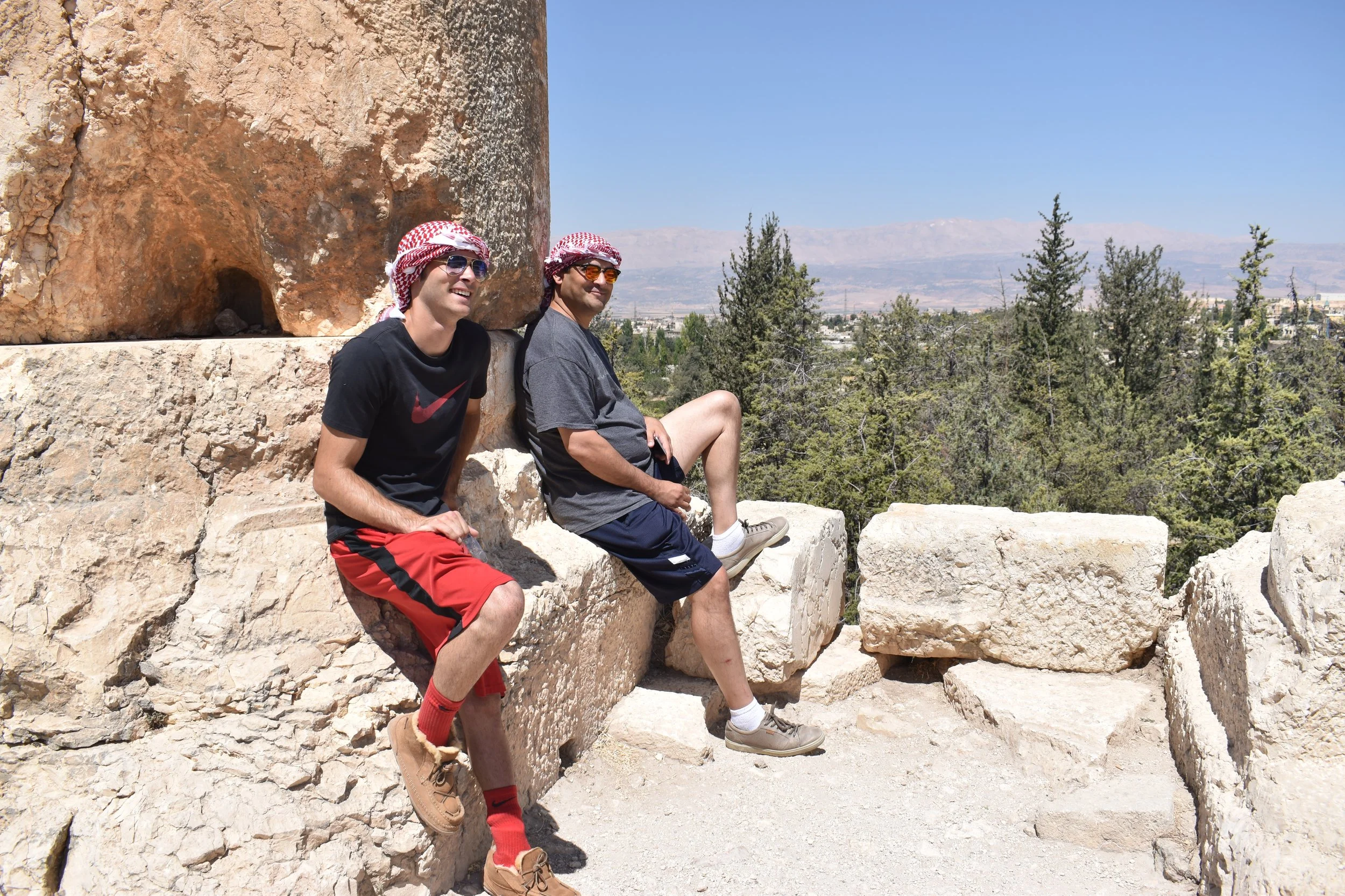 Two men sitting on large stone blocks in a desert landscape with trees and mountains in the background, wearing casual clothes, sunglasses, and headscarves.