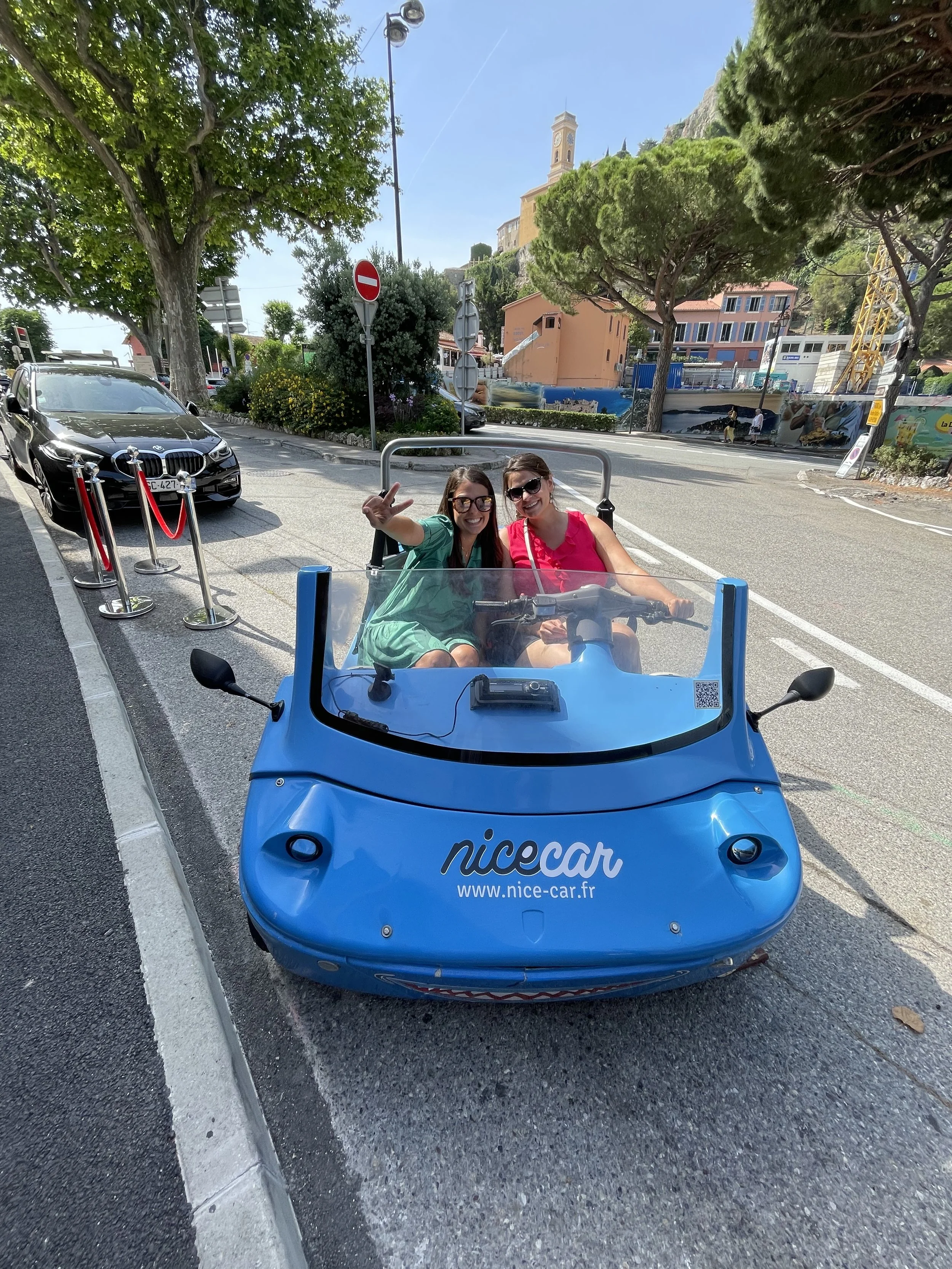Two women sitting in a blue electric scooter with the logo 'nicecar' and a website on it, smiling and posing for the camera, parked on a street with cars, trees, and buildings in the background.