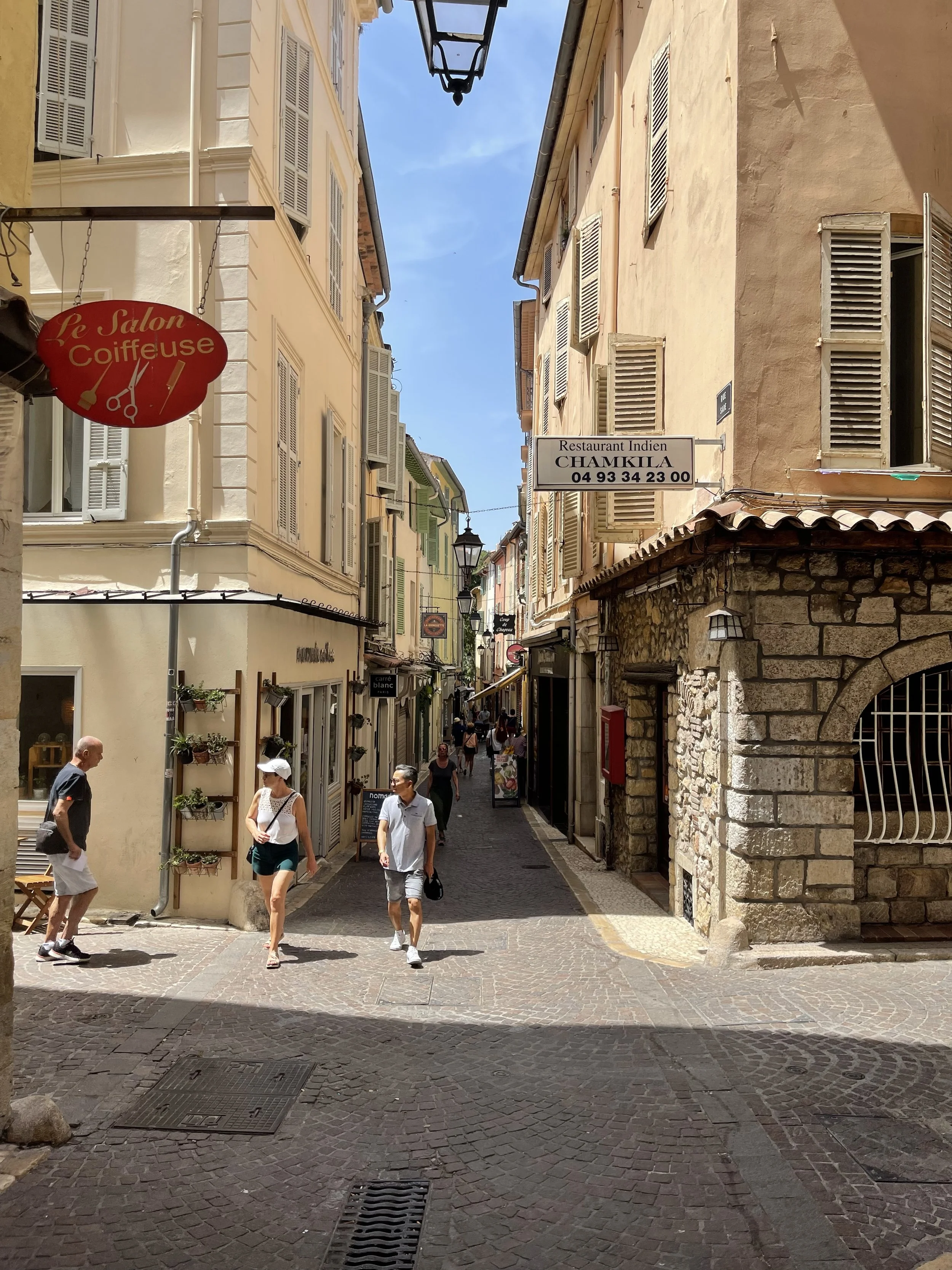 People walking along a narrow European street with colorful buildings and shops, bright blue sky.