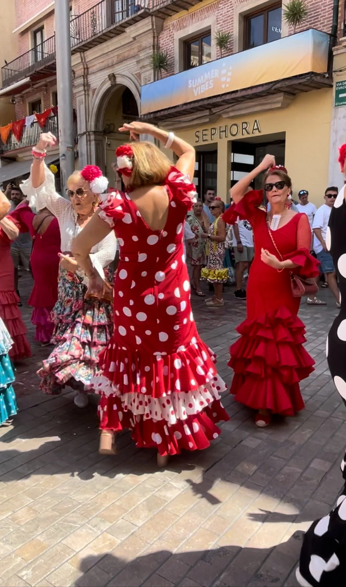People dressed in colorful flamenco dresses dancing on a street during a festival. The women are wearing red, white, and floral patterned dresses with ruffles. Some women have flowers in their hair. There are spectators in the background, buildings with balconies, and a sign that says 'SEPHORA'.