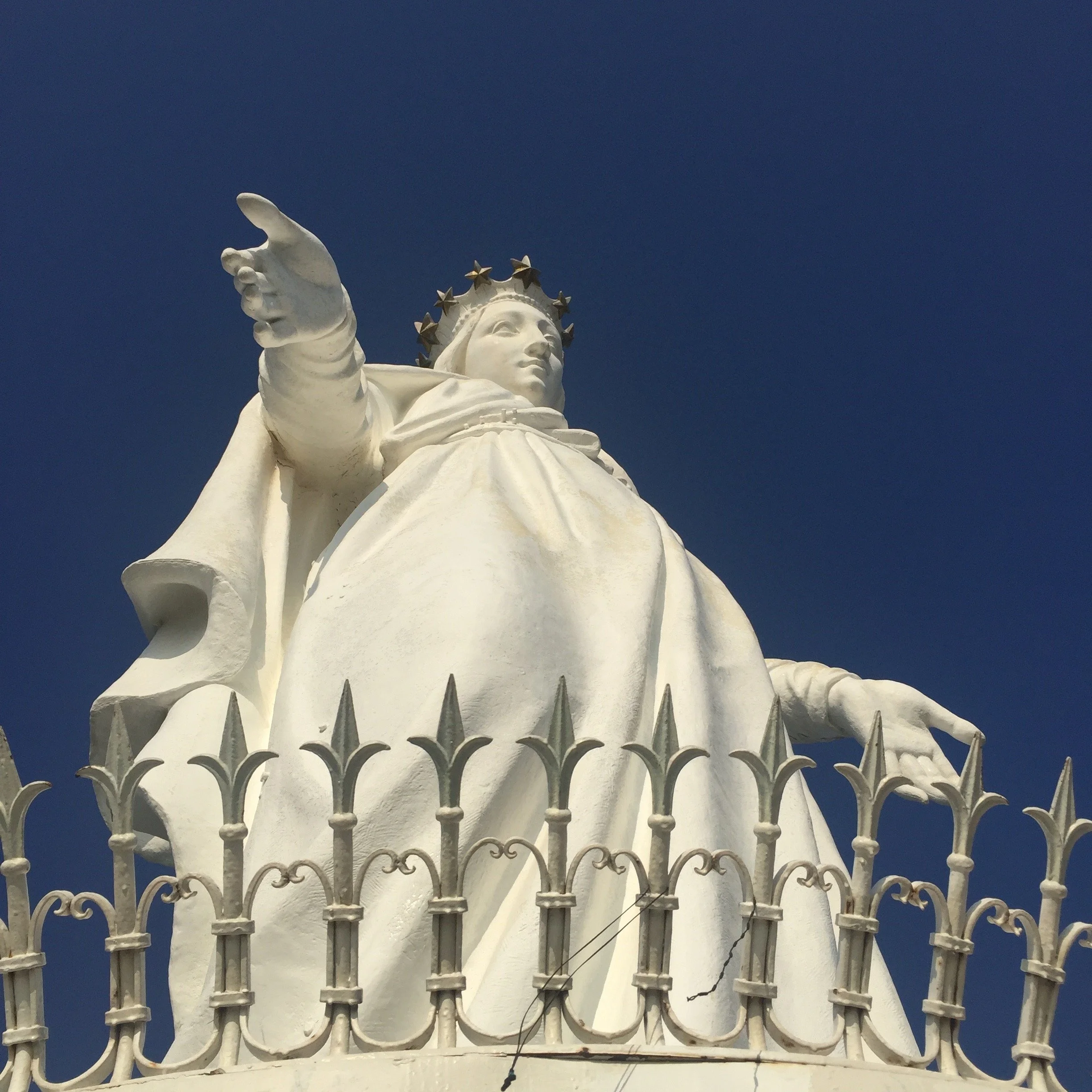 Close-up of a large white statue of the Virgin Mary with a crown of stars, reaching out with her right hand, against a blue sky, surrounded by a decorative white fence.