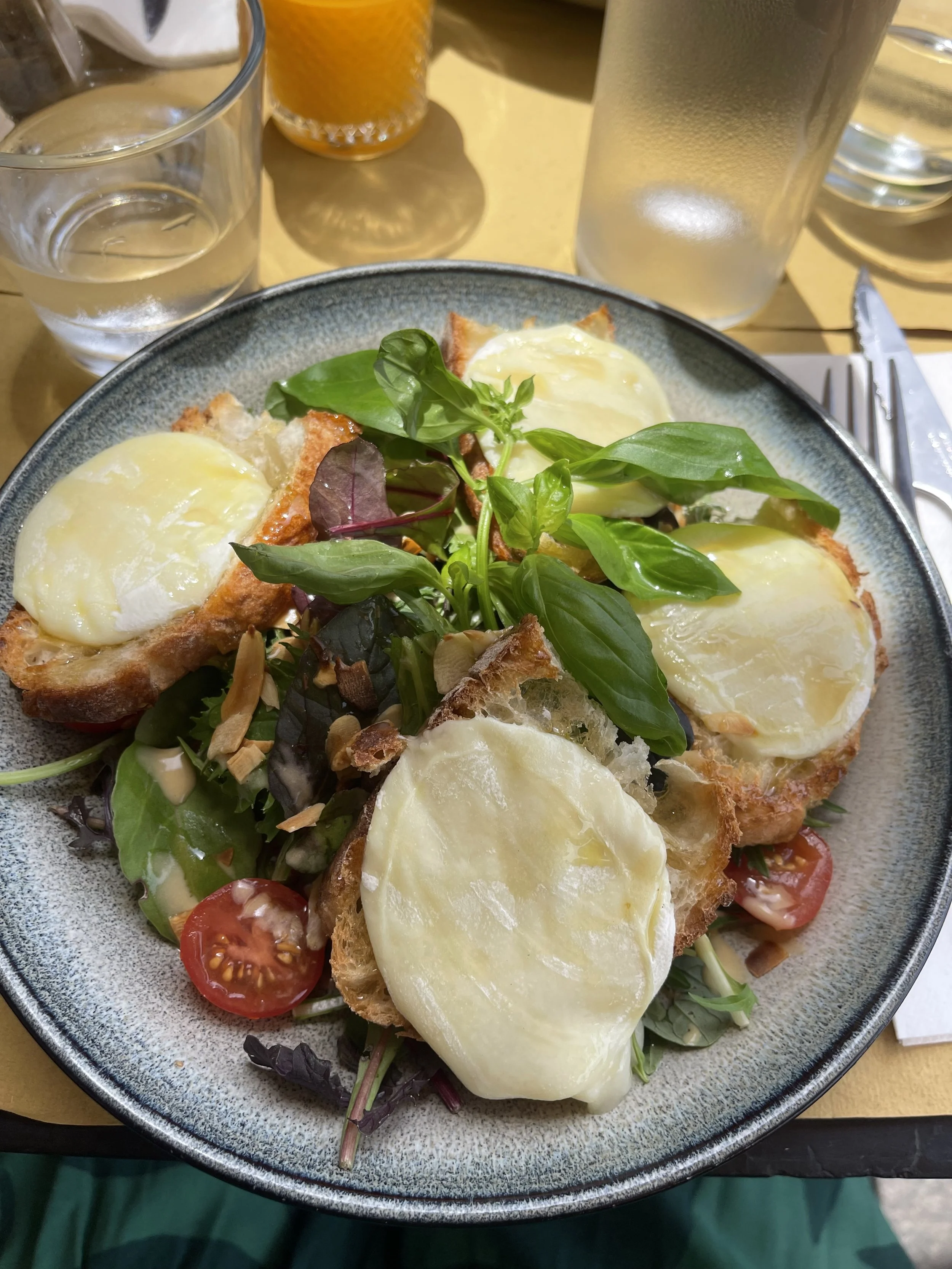 A plate of eggs Benedict with toasted bread, melted cheese, cherry tomatoes, mixed greens, and herbs, served on a table with a glass of water, a glass of orange juice, and utensils.