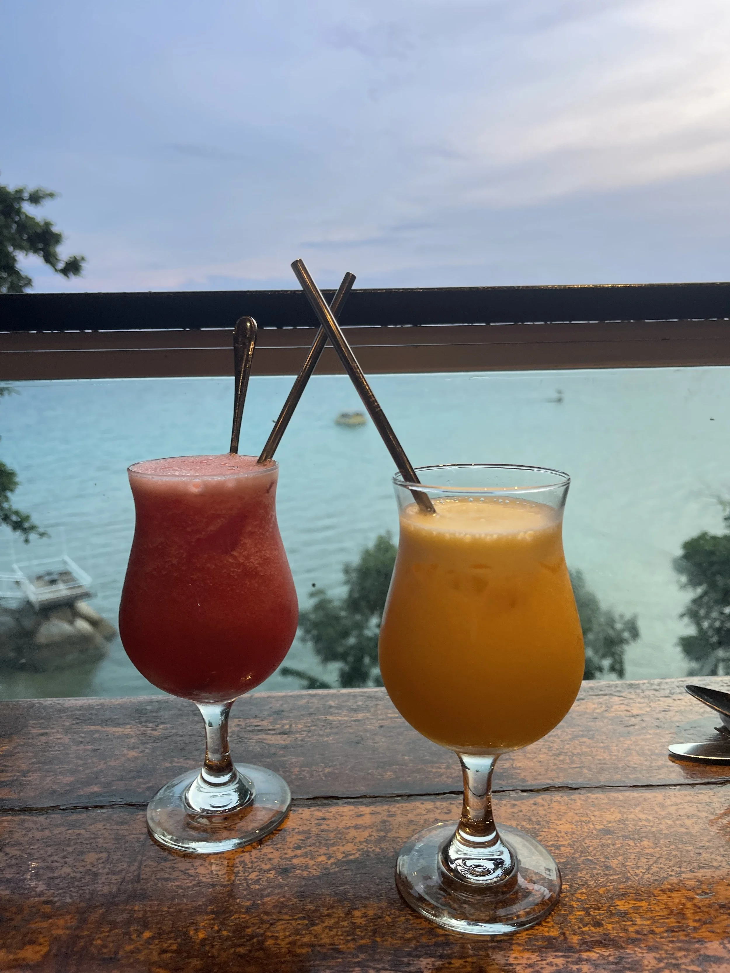 Two tropical drinks, a pink frozen cocktail and an orange juice, sit on a wooden table overlooking a body of water with boats and trees in the background.