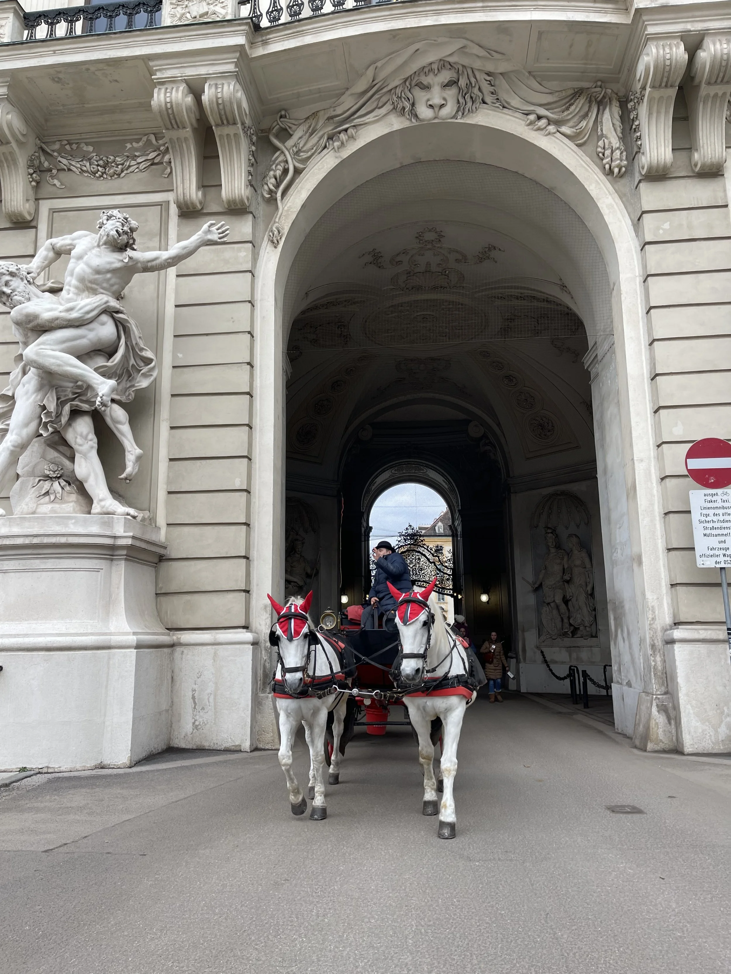 A horse-drawn carriage with a man riding, entering a large, ornate archway of a historic building with sculptures and decorative elements, including a lion's head above the arch.