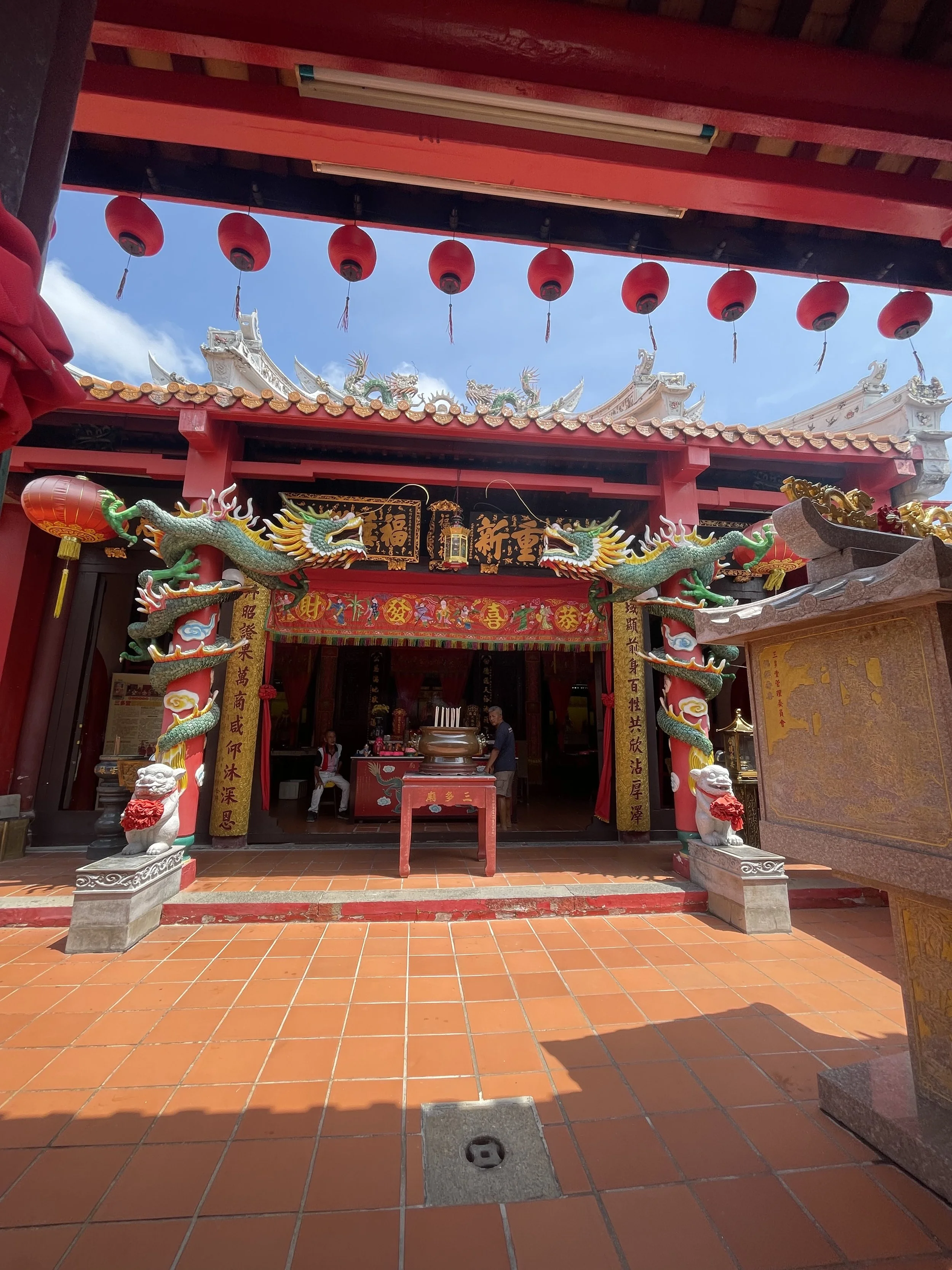 Entrance to a traditional Chinese temple with red pillars, dragon sculptures, and hanging red lanterns under a rooftop with ornate decorations.