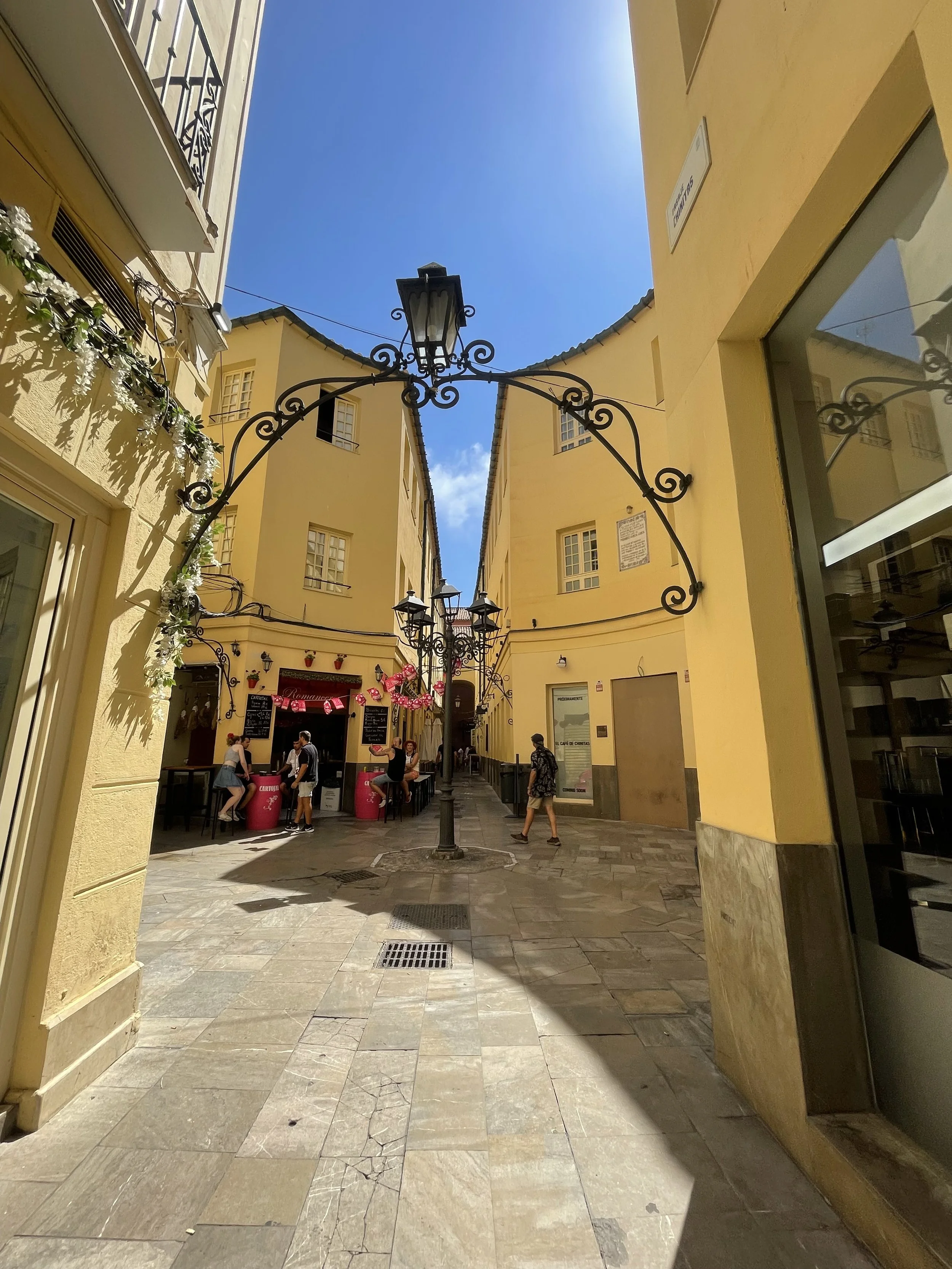 A narrow European-style street with yellow buildings, black ornate street lamps, and a blue sky. Pedestrians walk along the sidewalk, and a small cafe with pink decorations is visible in the background.