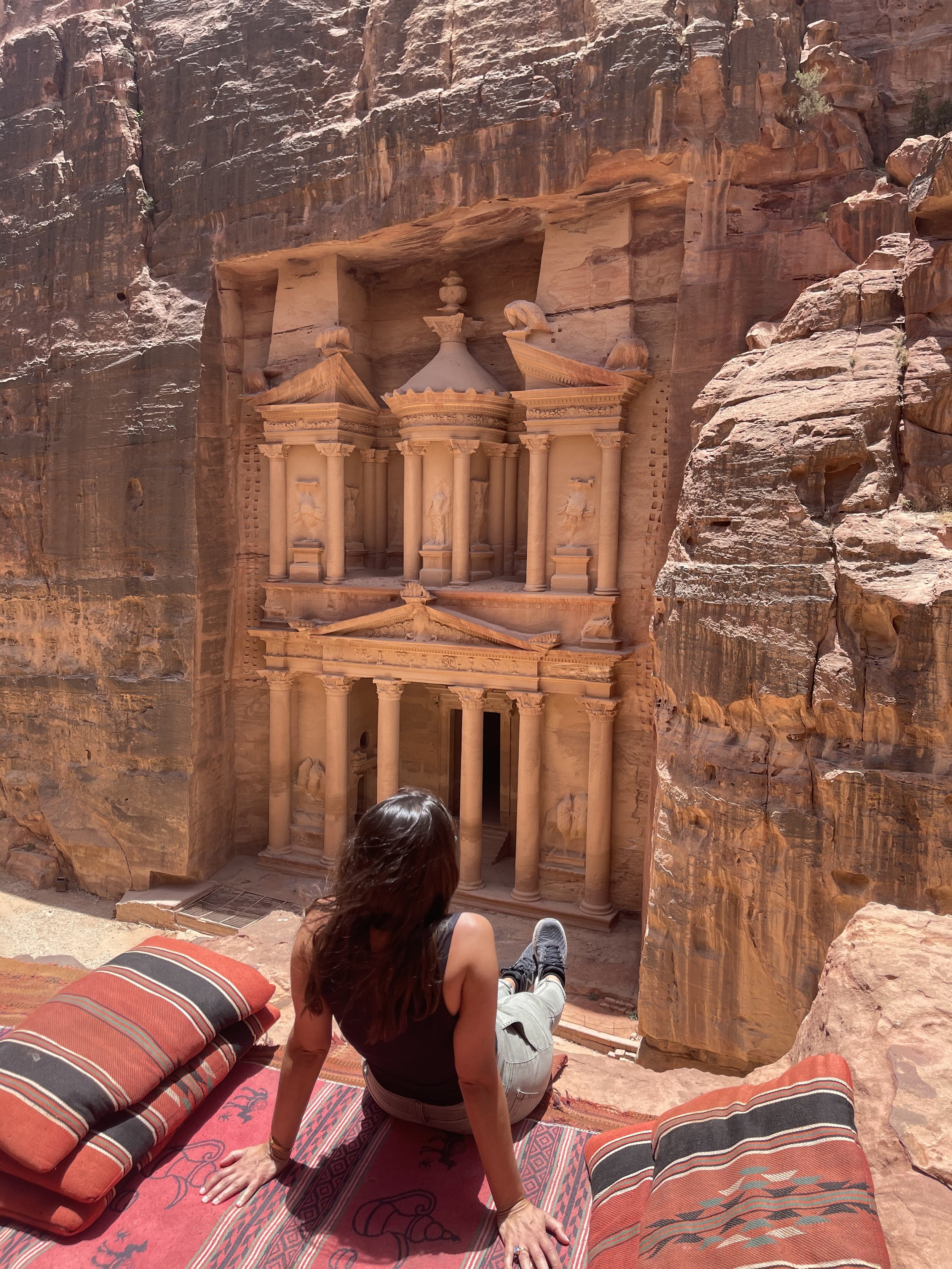 A woman sitting on a rug and cushions, overlooking the ancient rock-cut Treasury of Petra in Jordan, built into the sandstone cliff.