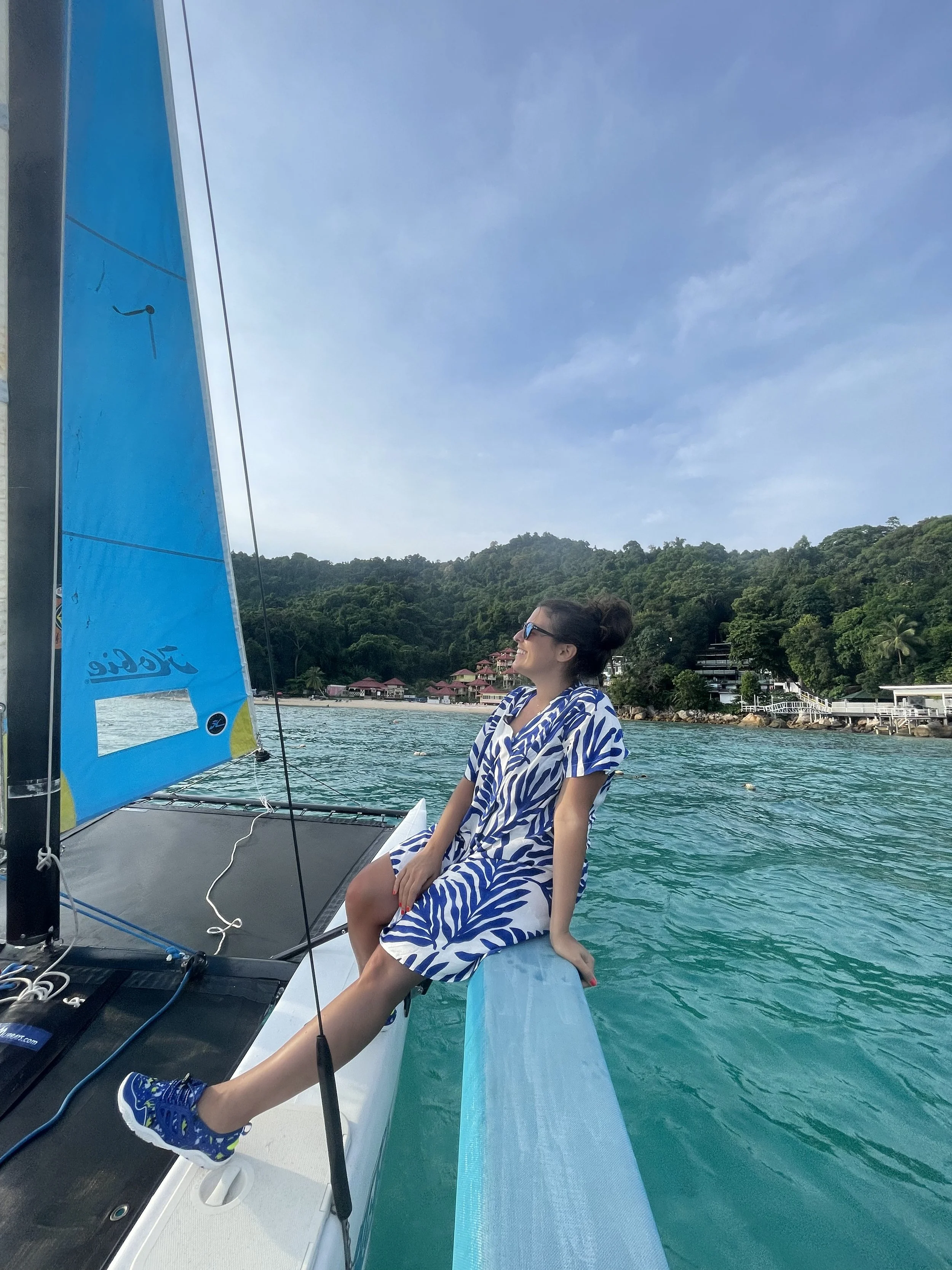 Woman in sunglasses sitting on the edge of a sailboat, wearing a blue and white patterned dress, with a tropical island and green hills in the background.