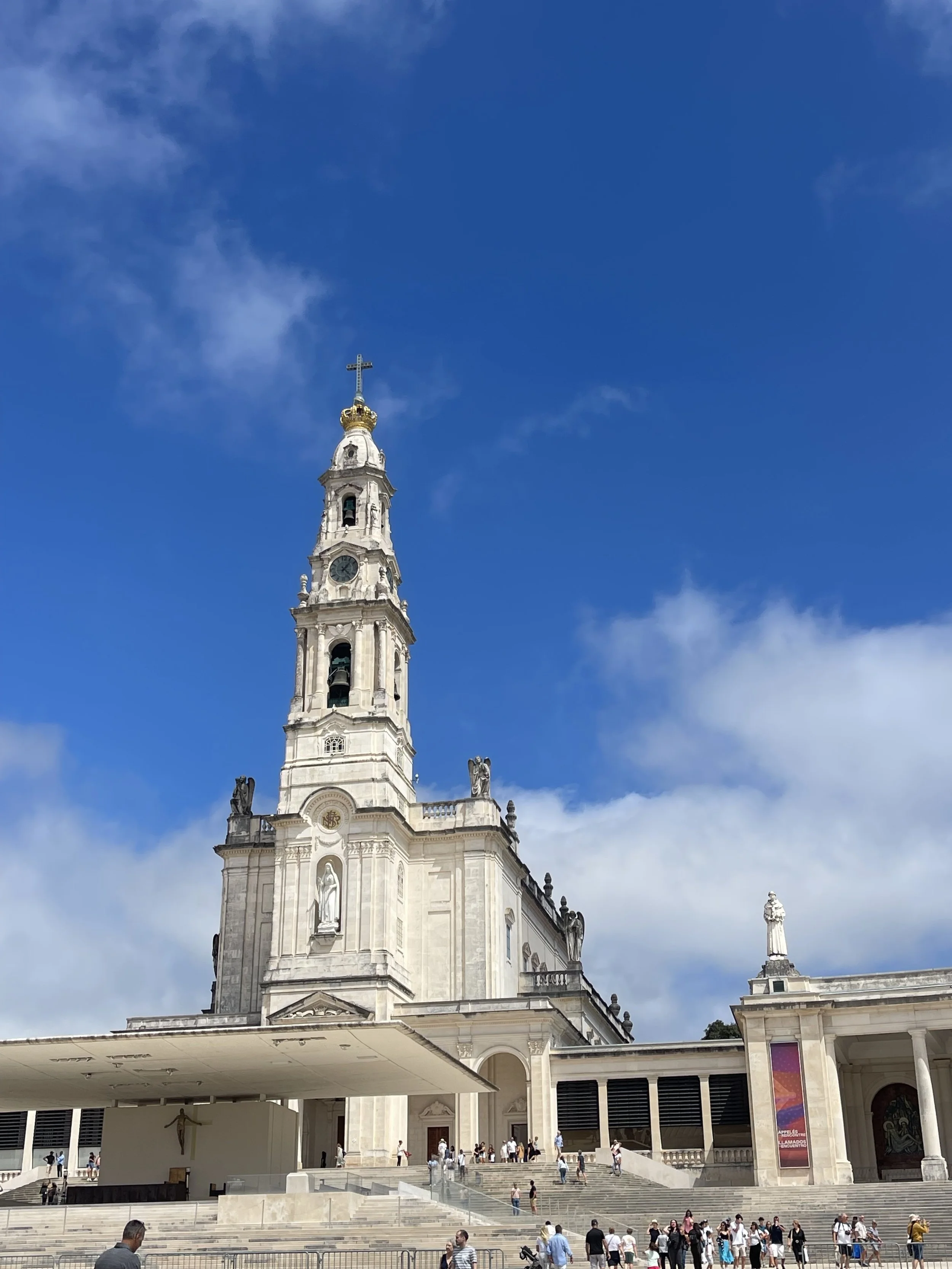The Basilica of Our Lady of Guadalupe, a large white church with a tall bell tower, numerous stairs leading to the entrance, and a crowd of people gathered outside on a sunny day with a blue sky and some clouds.