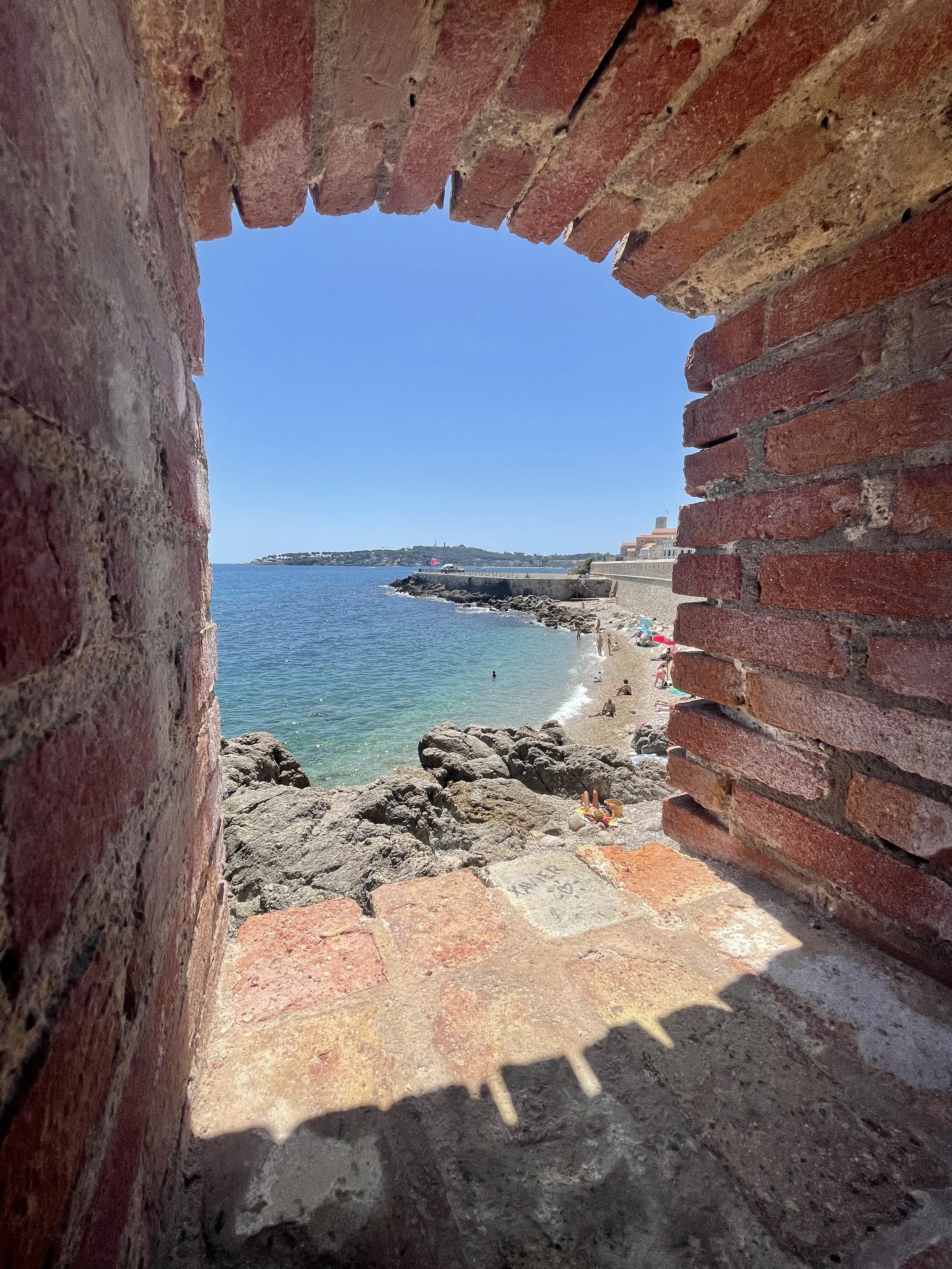 View of a beach seen through an old brick archway with rocks and people swimming and relaxing on the shore.