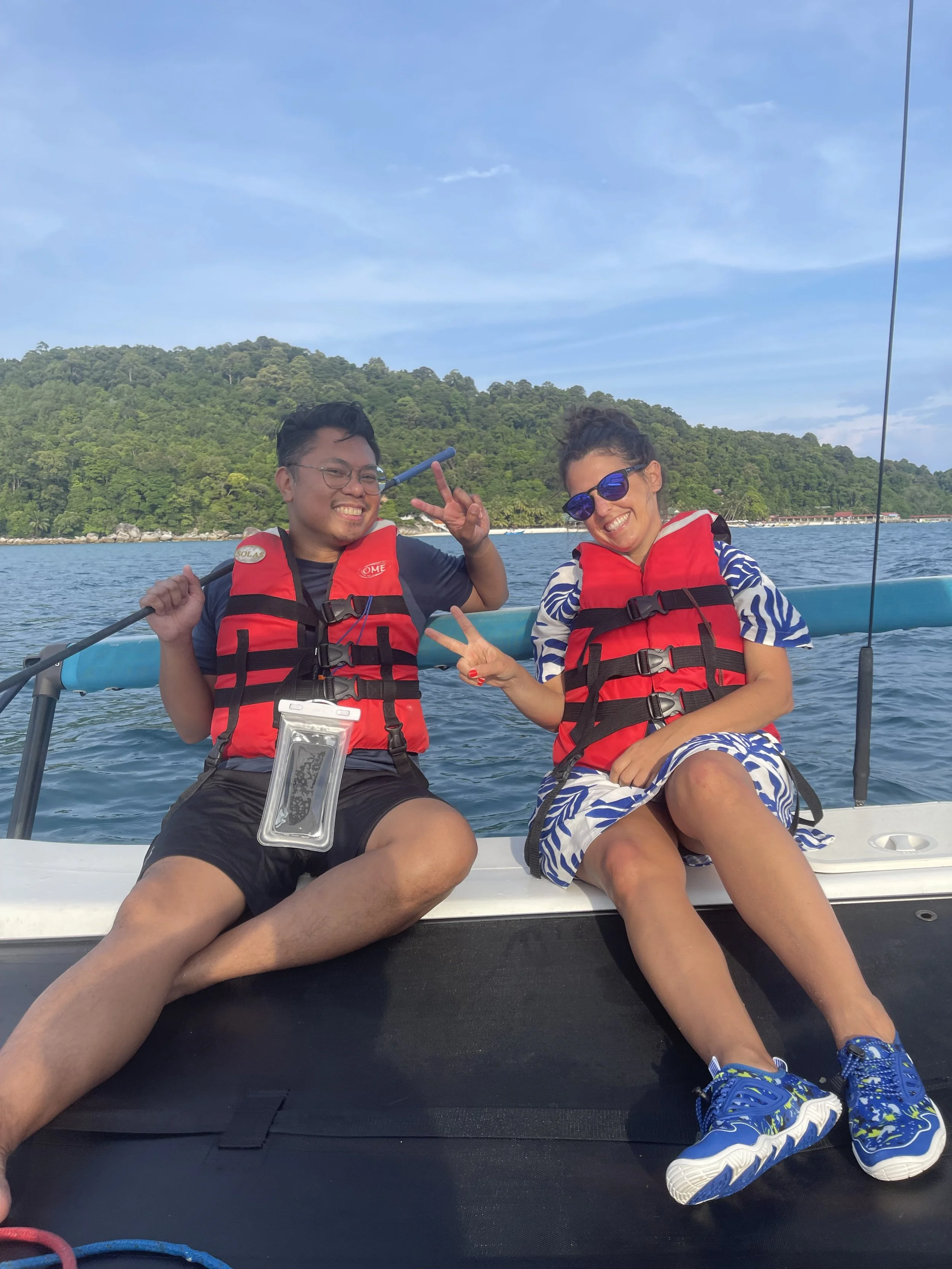 Two people sitting on a boat wearing life jackets, smiling, making peace signs, with a scenic background of water, forested hill, and blue sky.