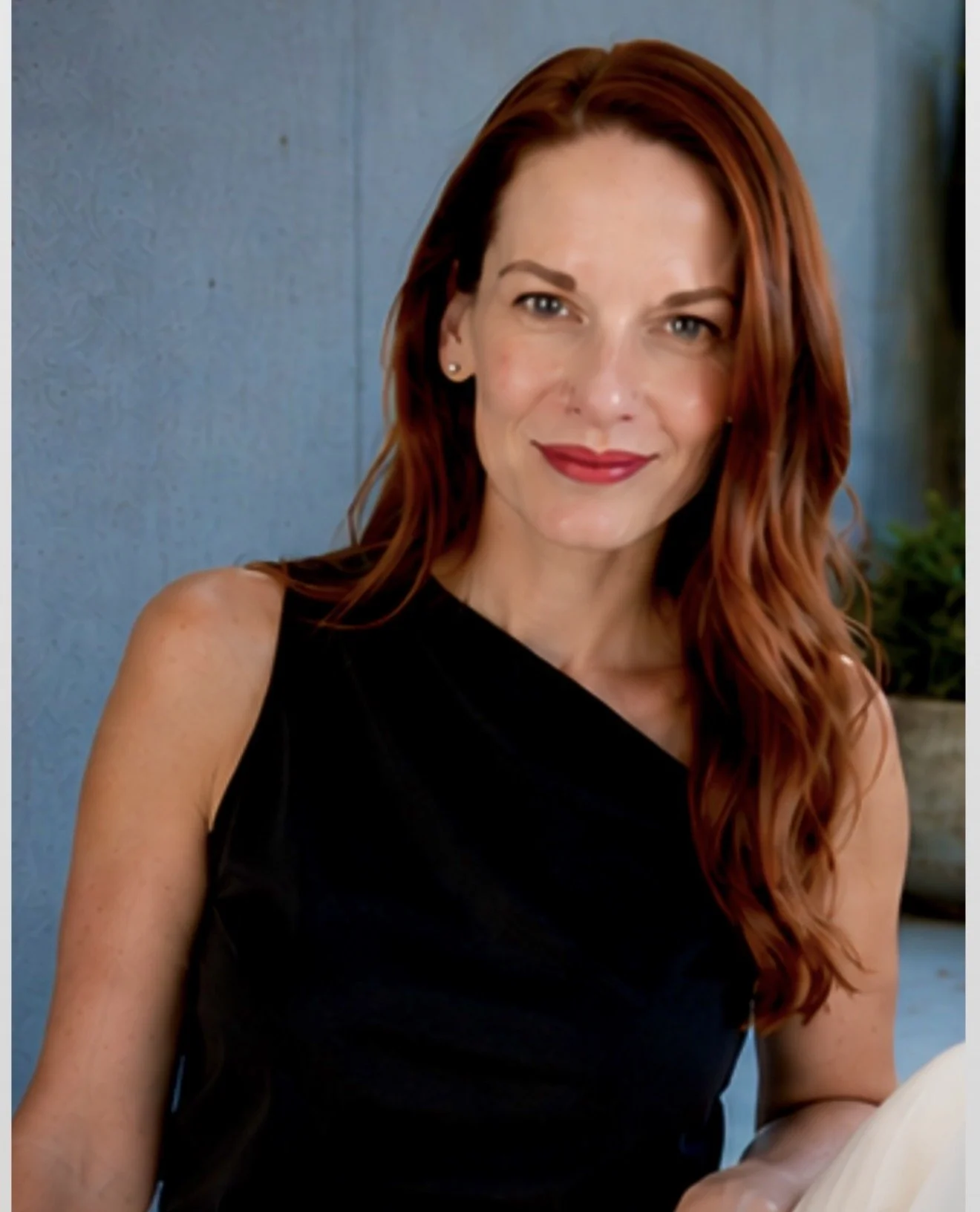 A woman with long red hair and fair skin wearing a sleeveless black top, smiling at the camera, standing against a blue wall background.