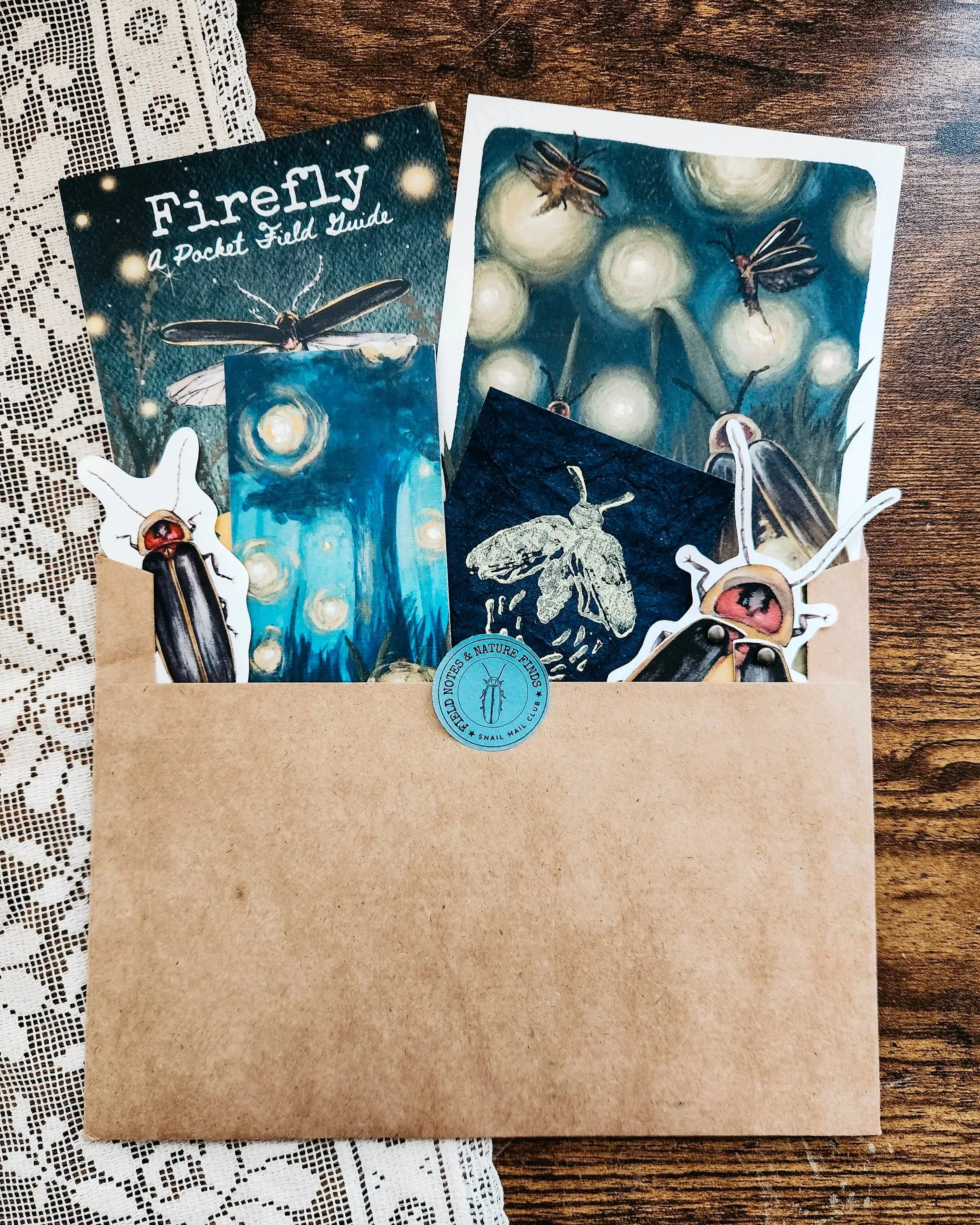 Firefly-themed snail mail nature study with illustrated paper goods, art prints, field guide, sticker, and handmade keepsakes arranged around an open envelope.