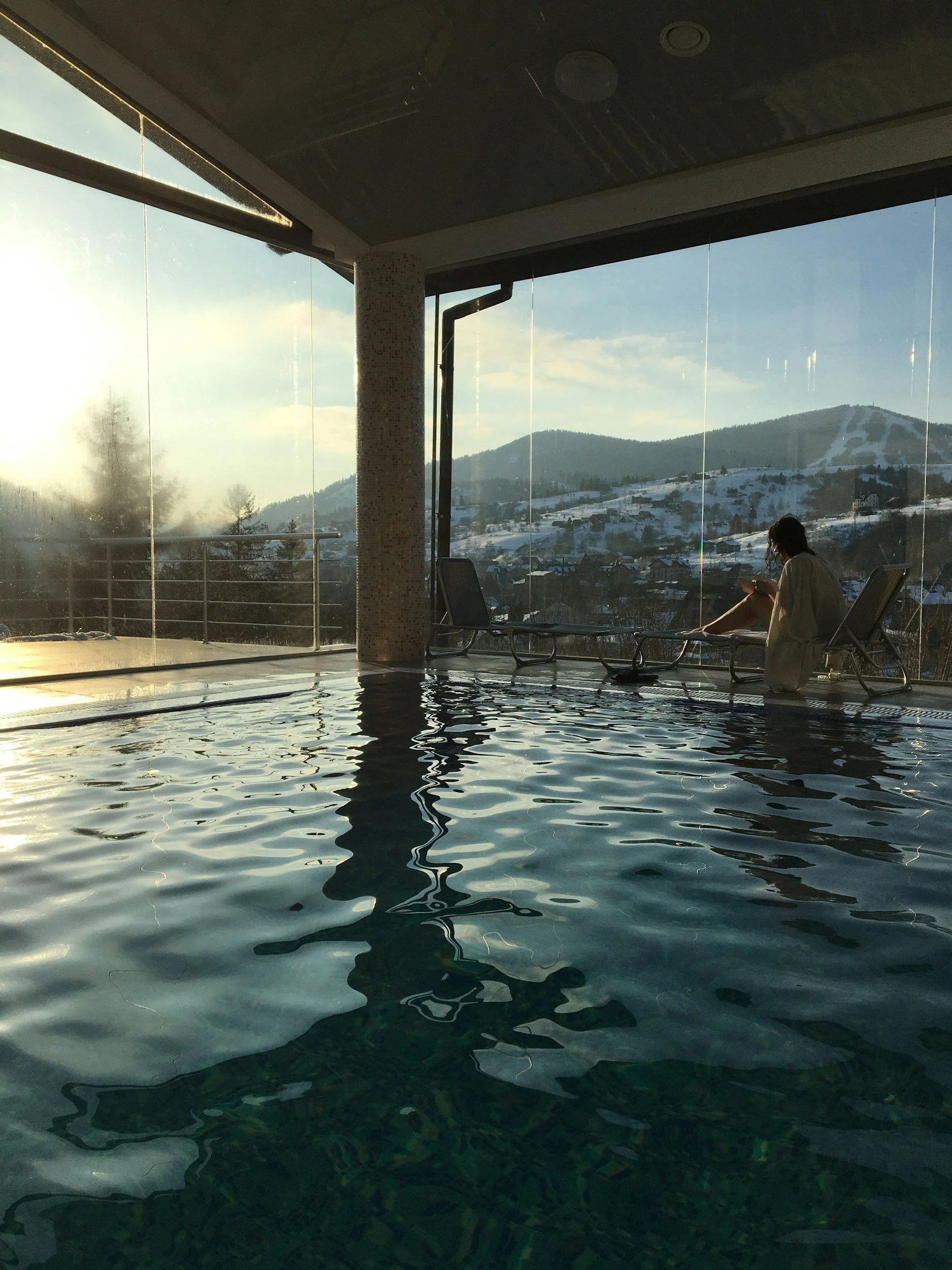 Indoor swimming pool with large windows showing snow-covered mountains and hills outside, a woman sitting on a lounge chair reading a book, and sunlight streaming in.