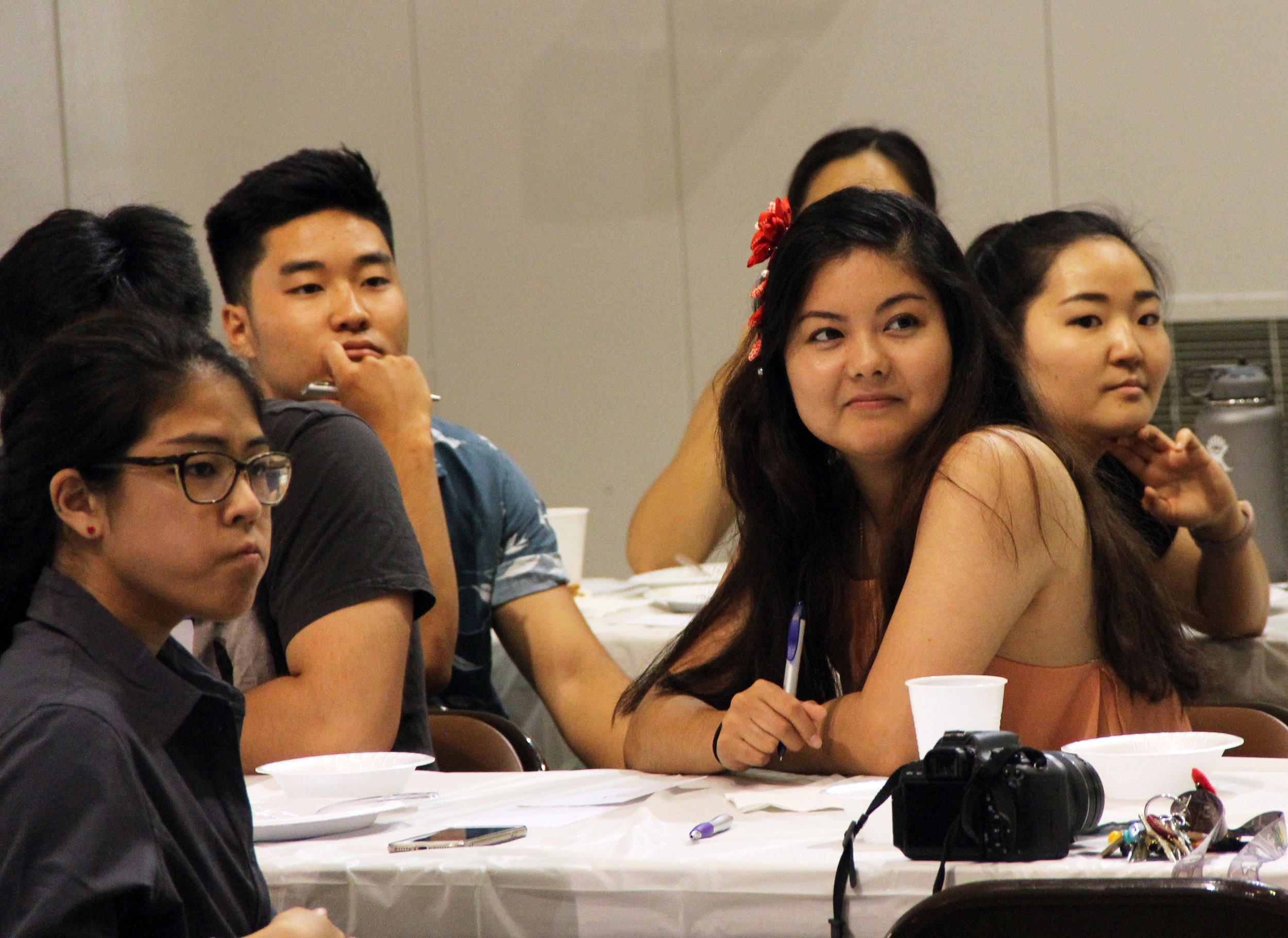 A woman holds a pen and listens to a speaker while at a table surrounded by other men and women.