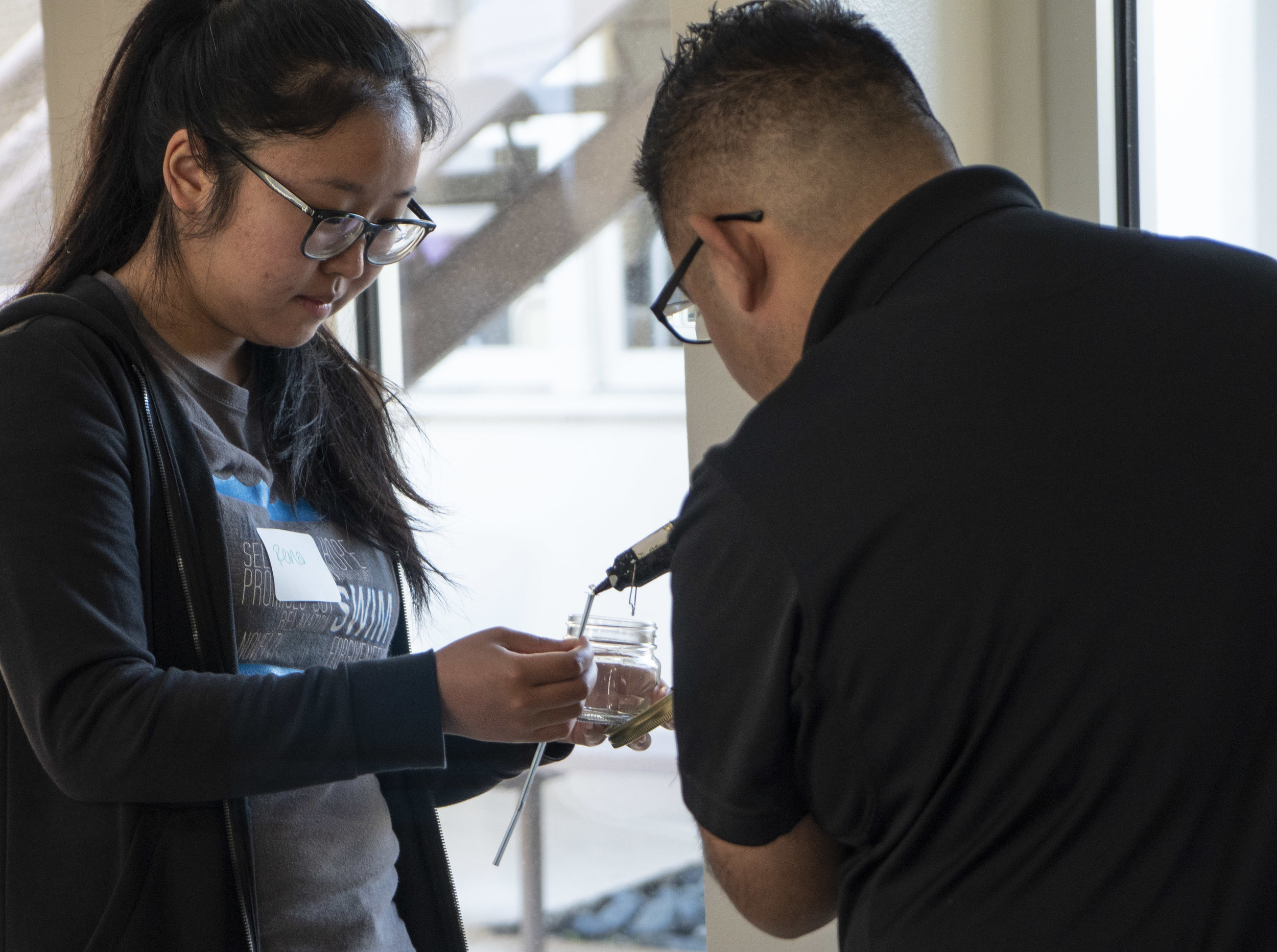 A man adds hot glue to the bottom of a wick for a woman wearing glasses who is making a candle. 