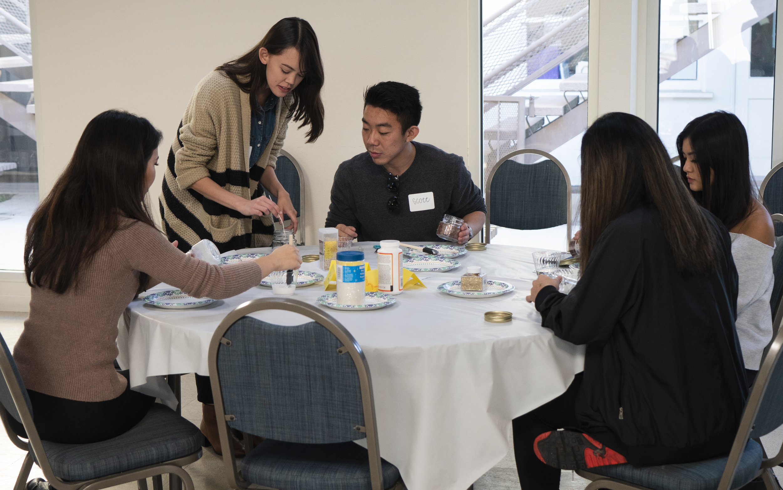 A group of women and a man sit at a round table decorating their glass containers.