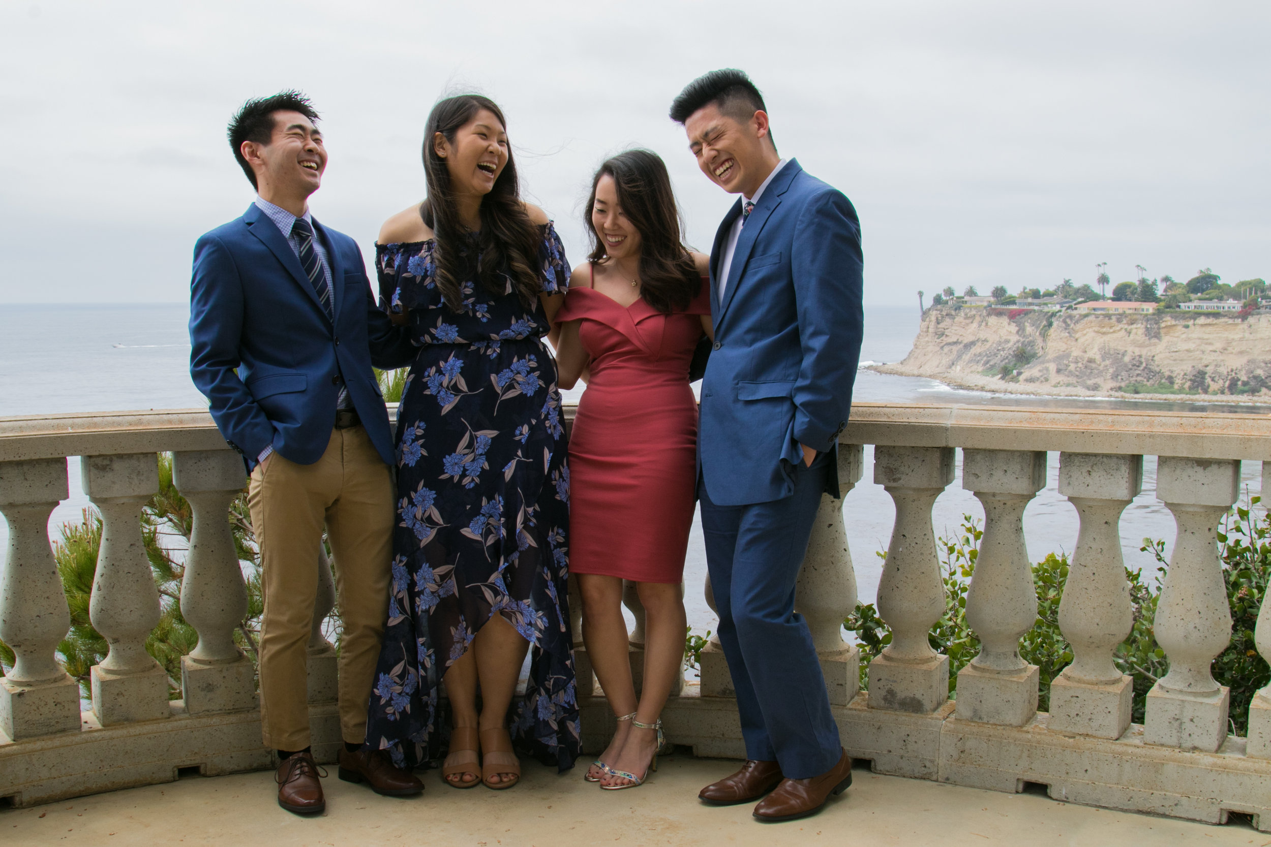 Two men and two women wearing suits and formal dresses laugh outside on a terrace overlooking water behind them.