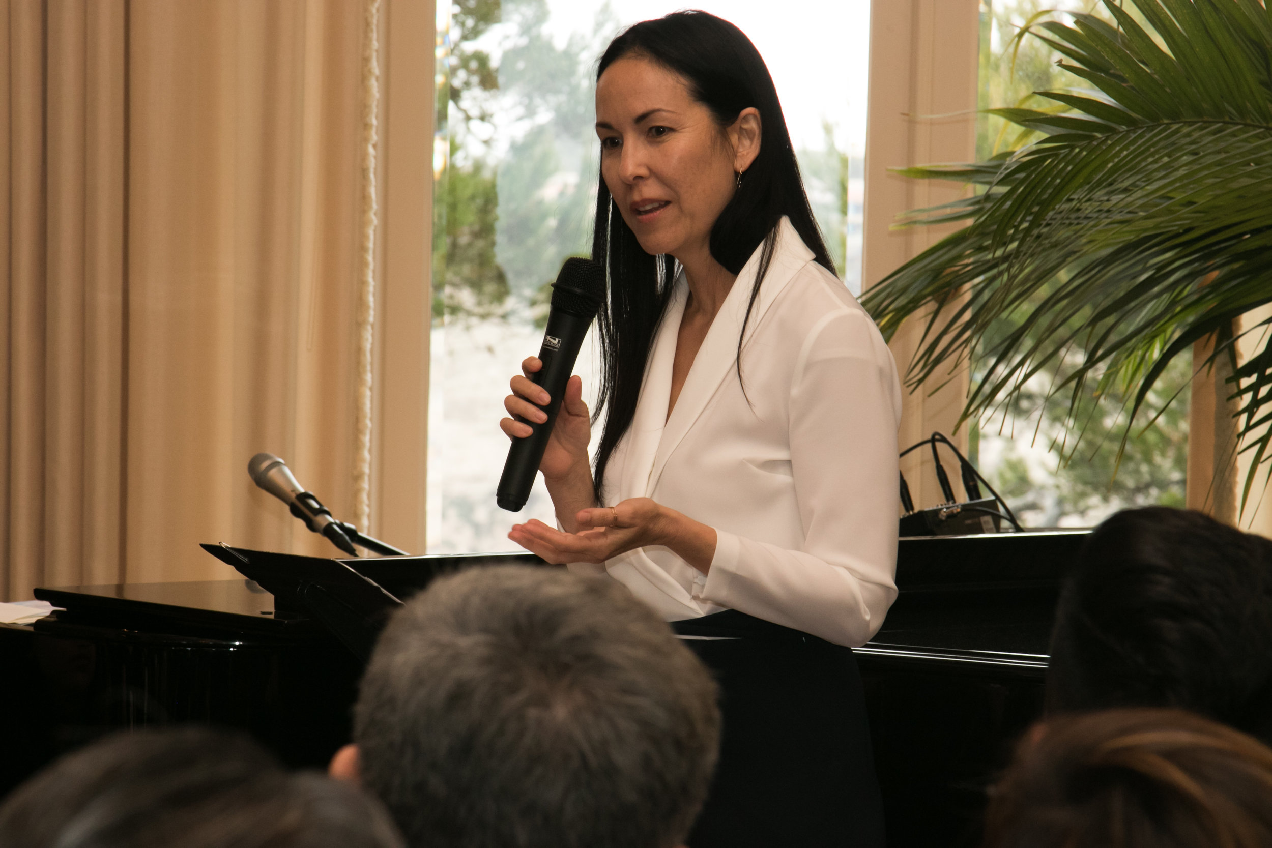 A woman with long straight black hair speaks with a microphone to a group with a piano behind her.