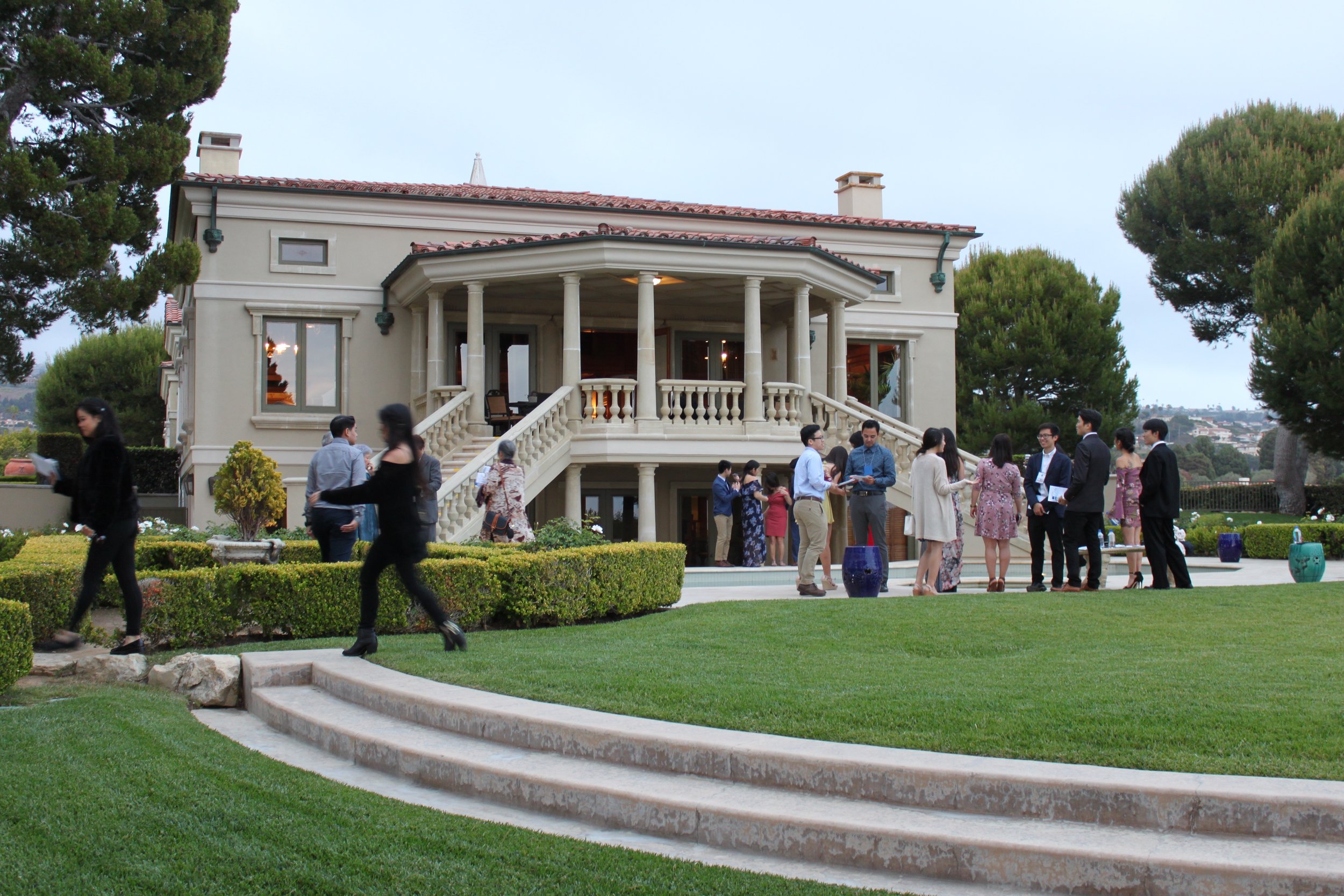 People gather on a lawn with staircases on either side of the building leading up to the terrace in the background.