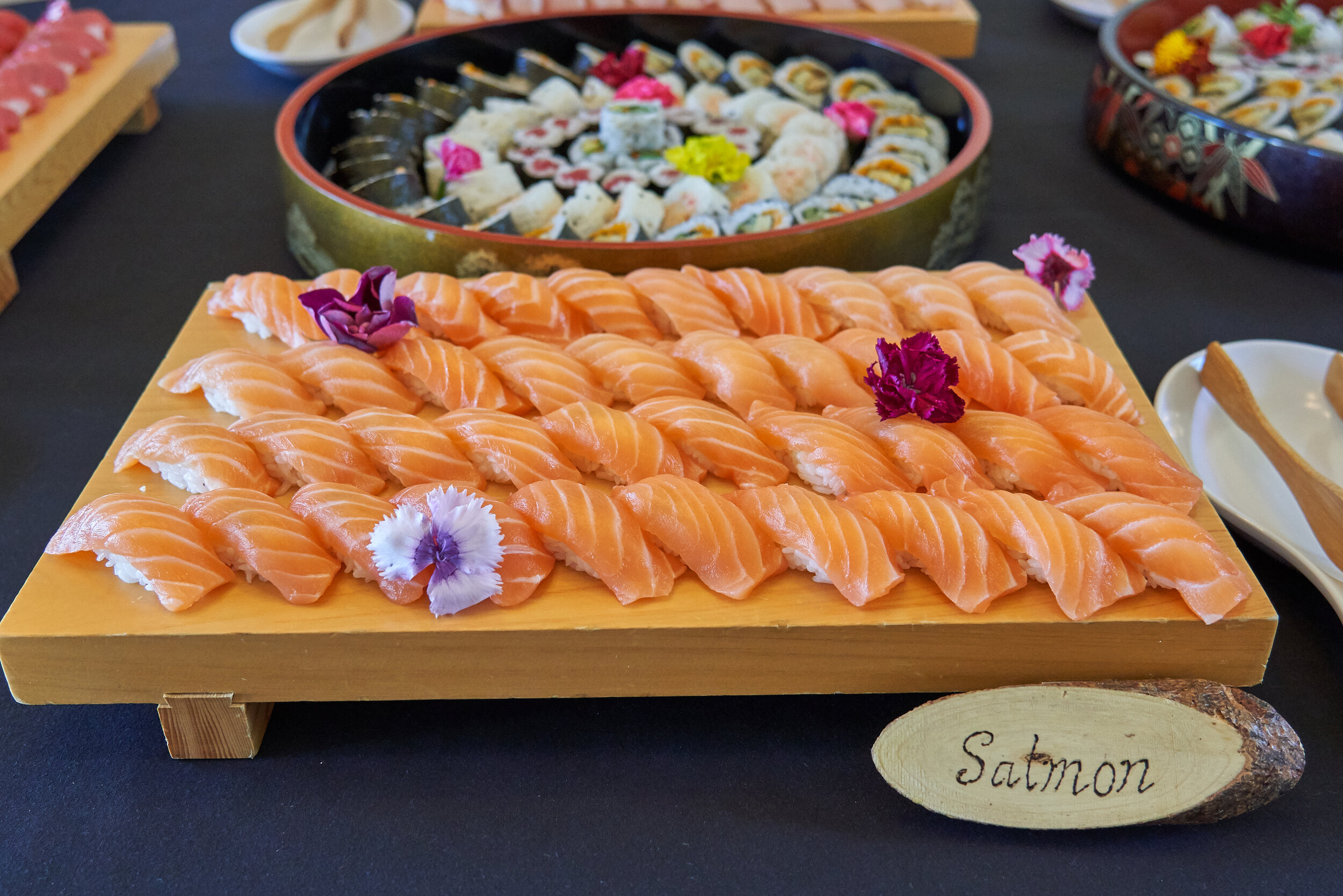 A large salmon sushi platter rests on a table with multi platters of sushi in the background.