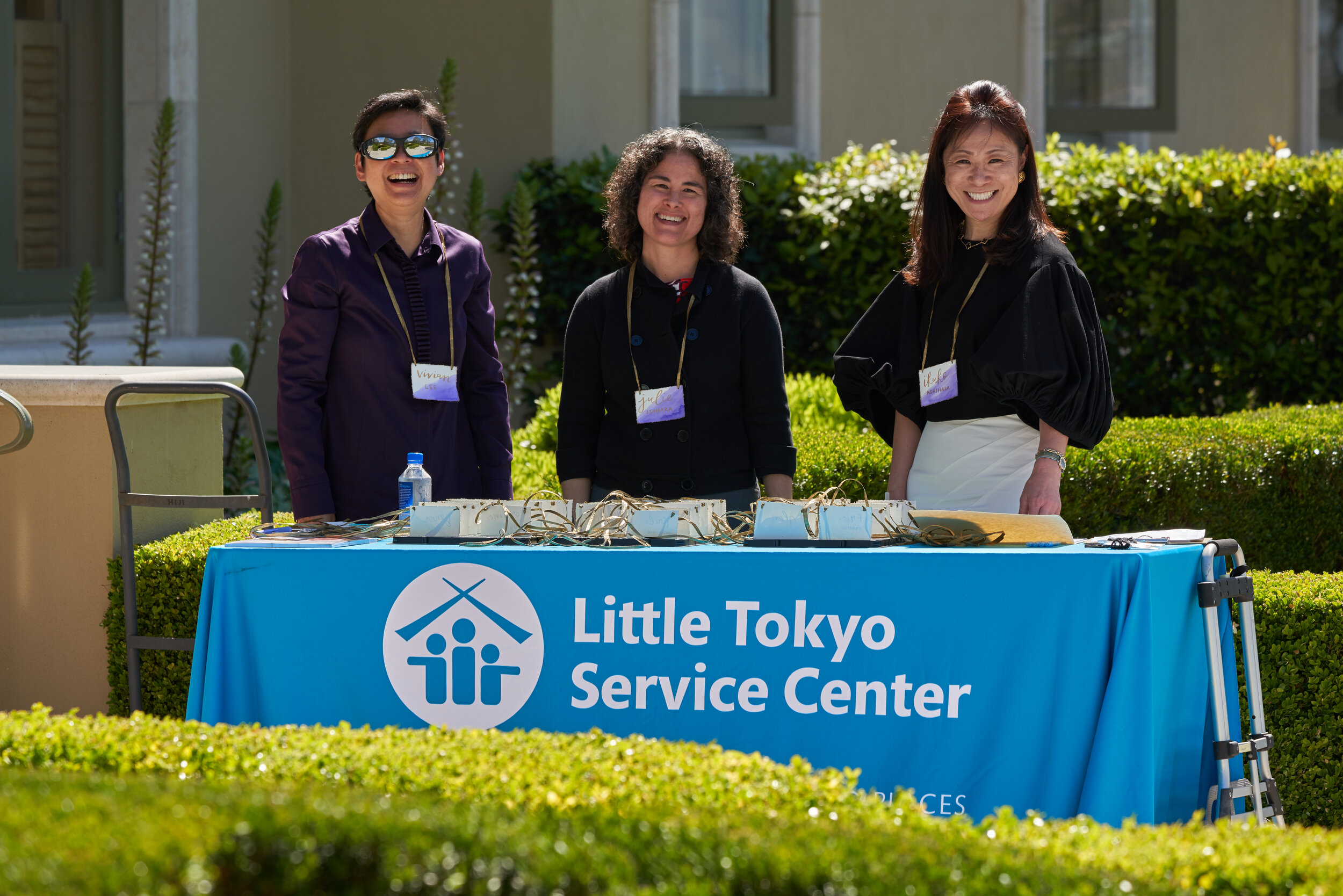 Three ladies smile behind a table with a cloth on it that reads “Little Tokyo Service Center.”