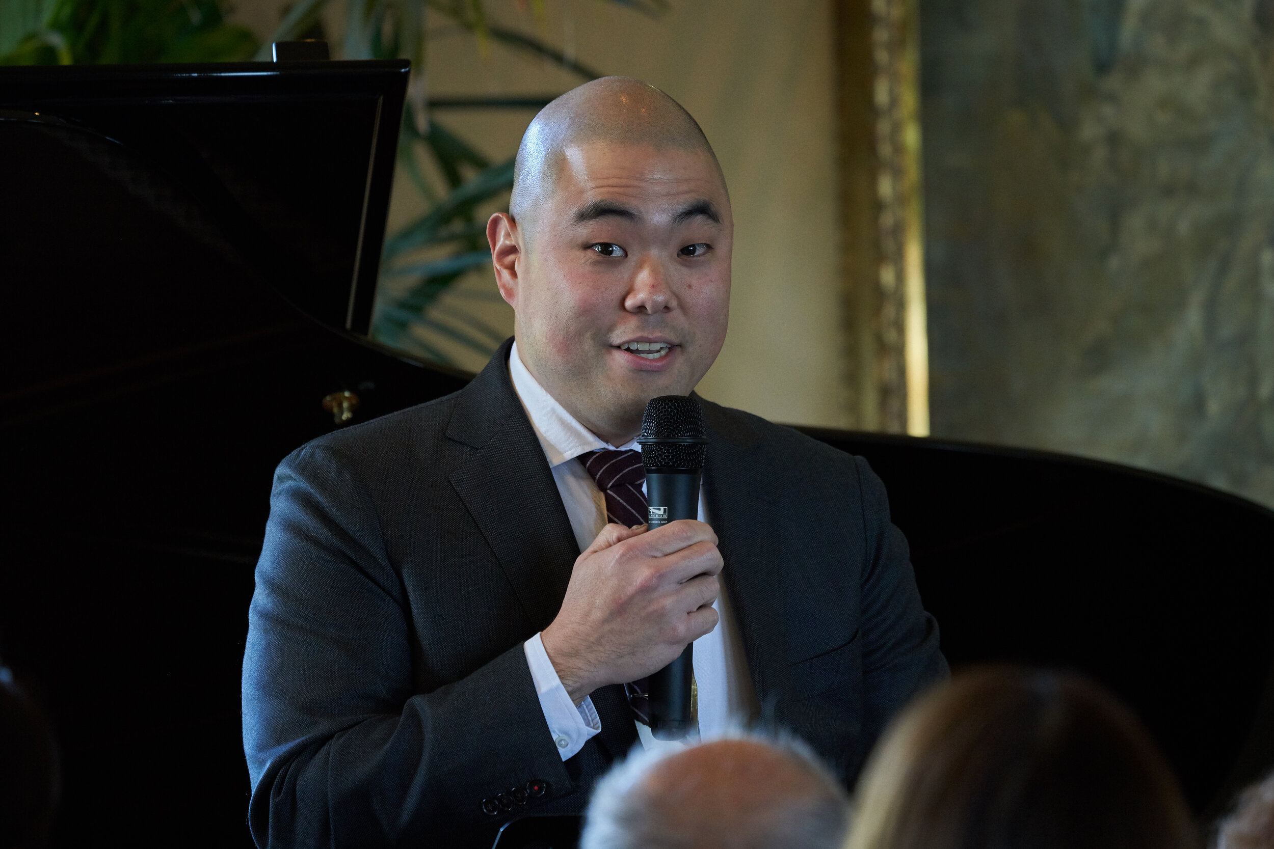 A man with a shaved head holds a microphone in front of a group of people with a piano behind him.