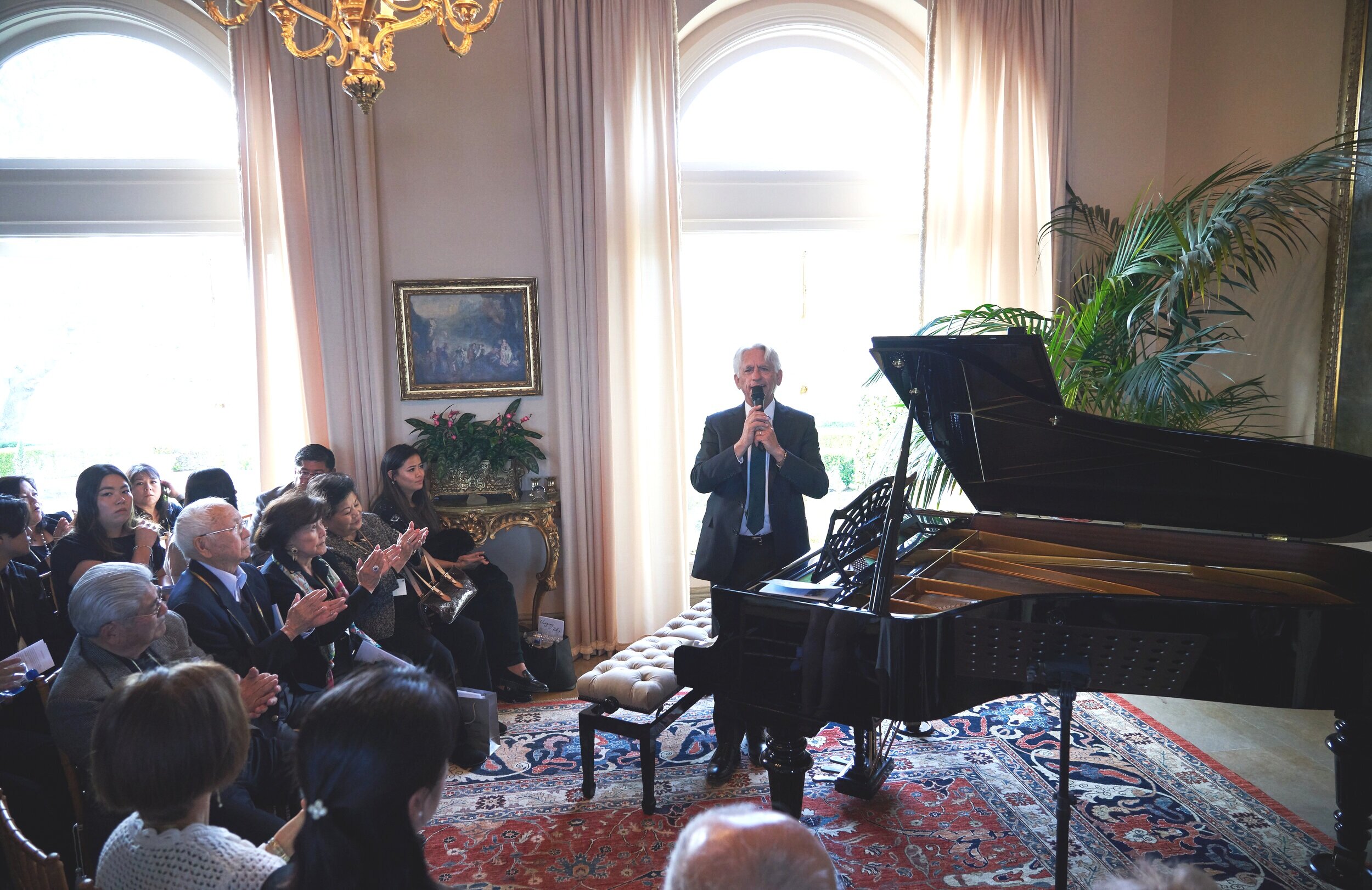 A man with white hair stands next to the piano as he speaks with a microphone to a seated group.