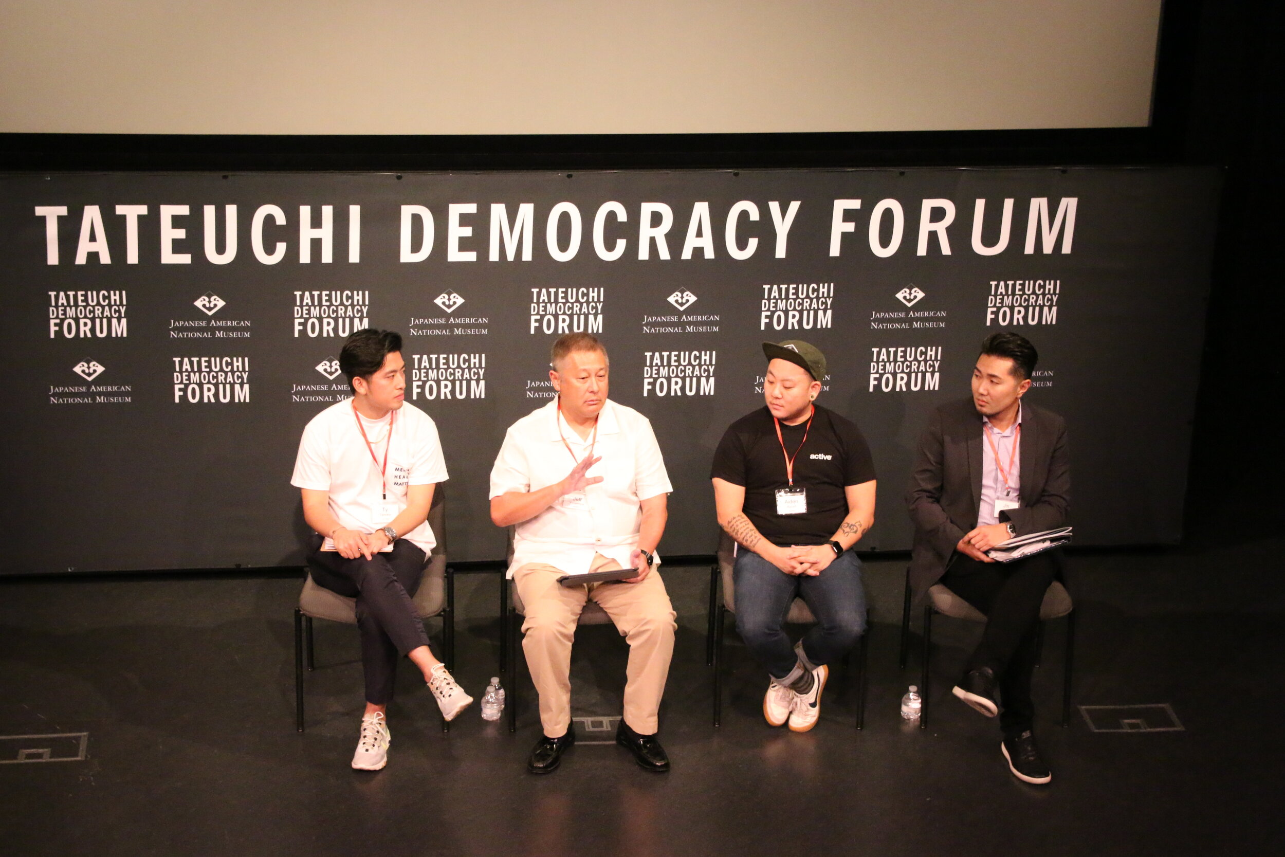 Four men sit in front of a step and repeat background that says “Japanese American National Museum” and “Tateuchi Democracy Forum.” 