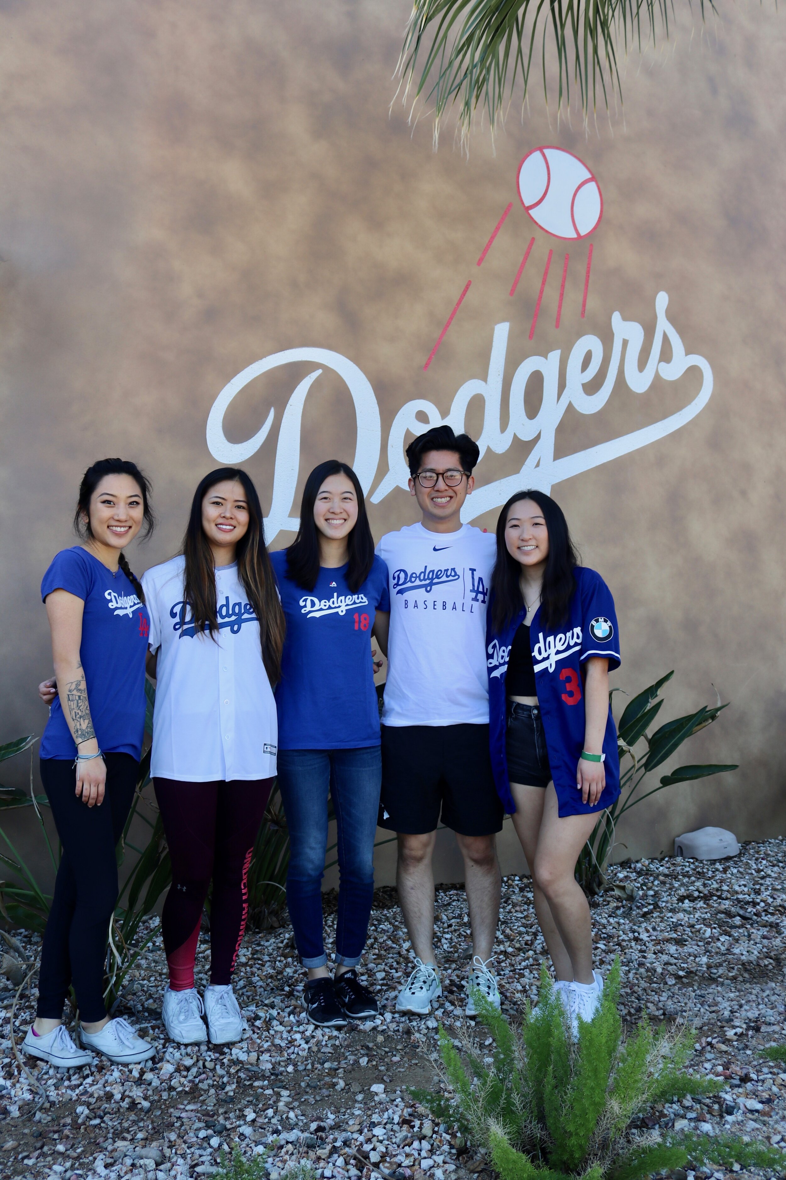 A group of men and women smile wearing Dodger’s shirts in front of the Dodger’s painted sign on a building. 