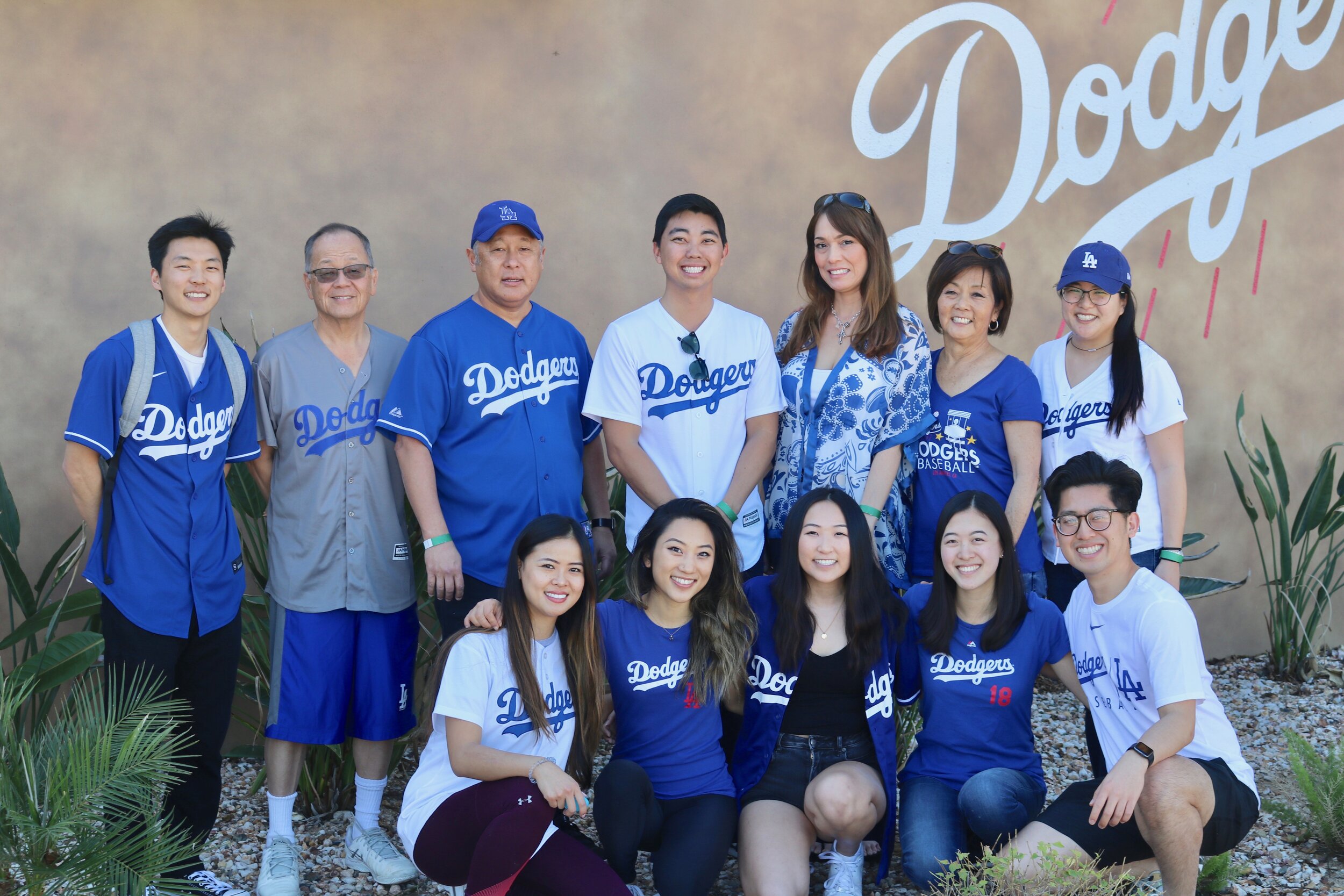 A large group of men and women smile wearing Dodger’s shirts in front of the Dodger’s painted sign on a building. 