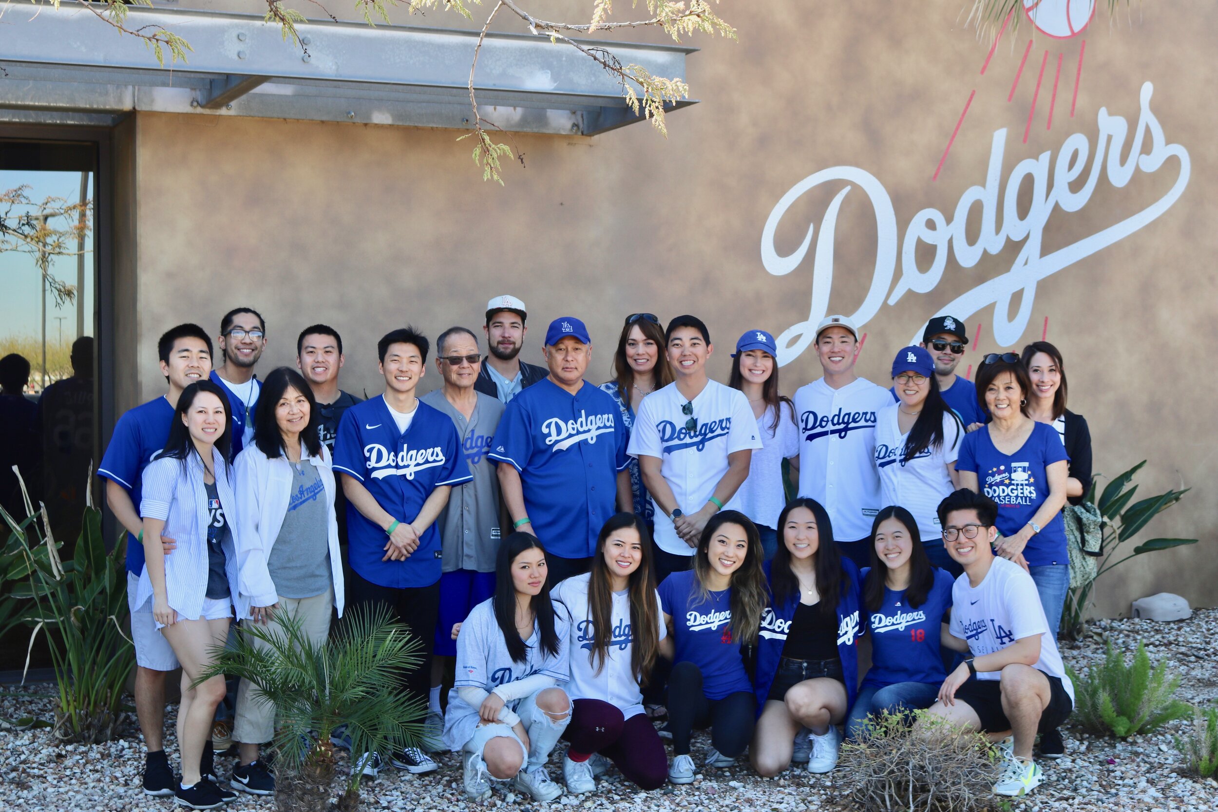 A large group of men and women smile wearing Dodger’s shirts in front of the Dodger’s painted sign on a building. 