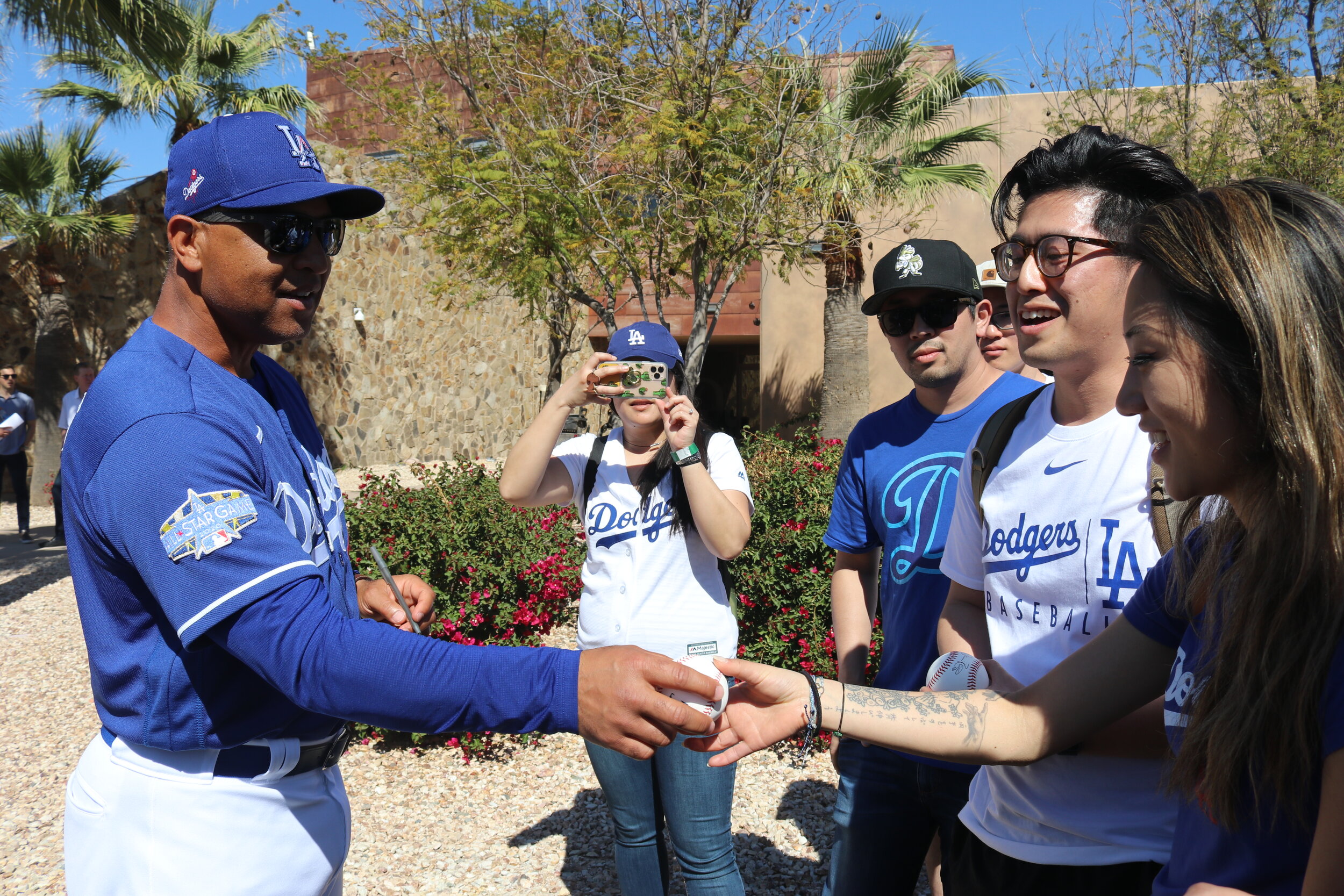 A man wearing a Dodgers jersey hands a fan a ball while another takes a photo.