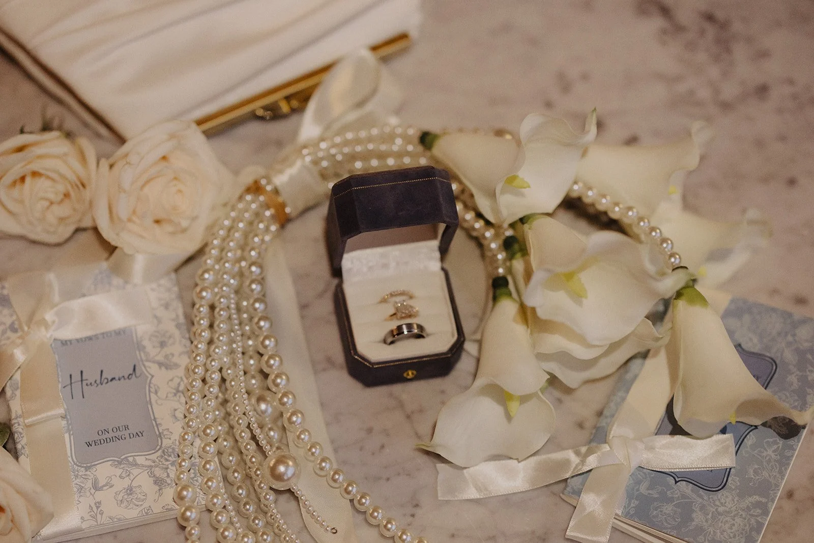 A wedding detail flat lay at Elm Lagrange displays blue + white vow books, wedding rings, and calla lillies