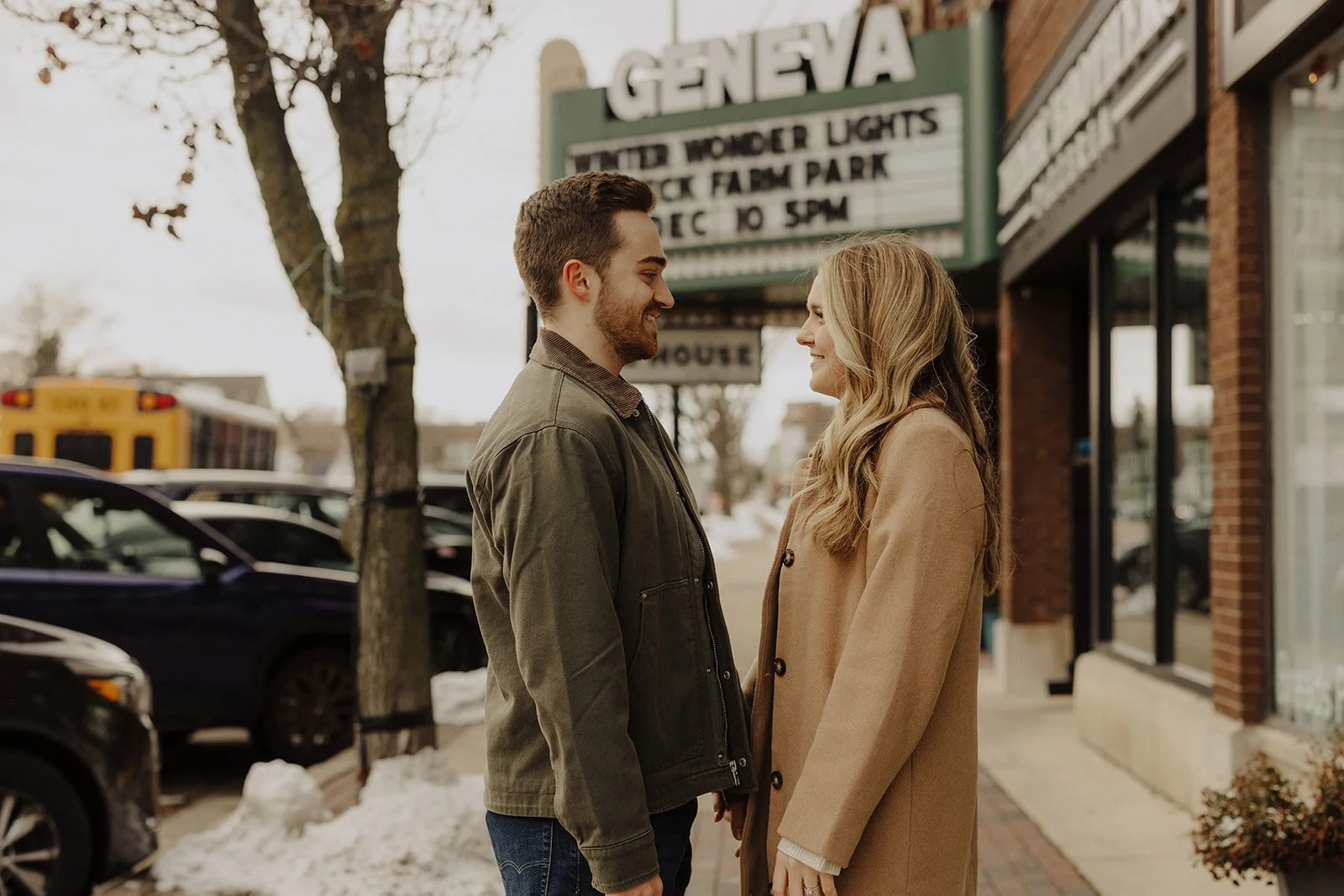 A couple smiles at each other outside Geneva Ale House during winter engagement photography Geneva, IL