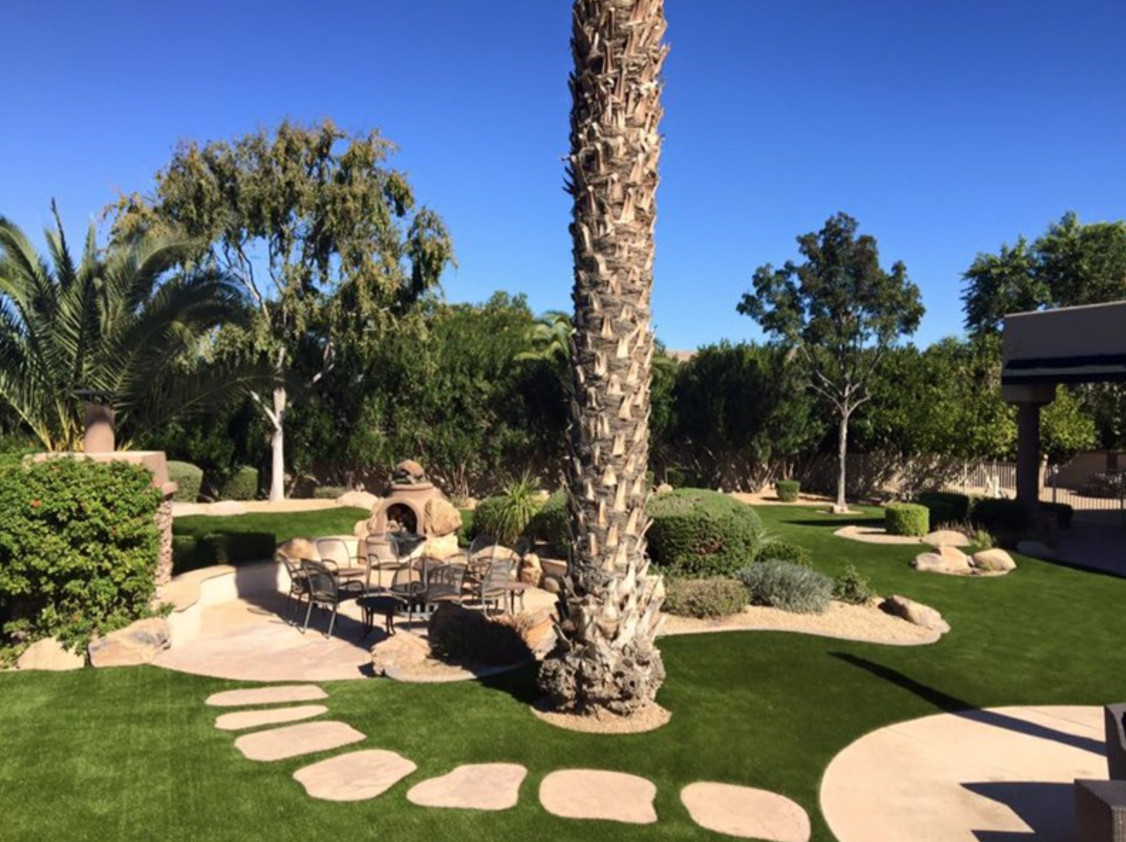  Artificial grass installation with concrete stepping stones inset surrounding paver patio with fire pit and palm trees. 