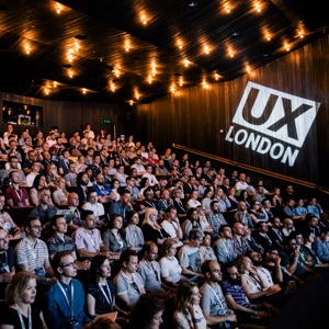 A photo of a many people sitting in a large auditorium with a sign behind them that reads, “UX London.”